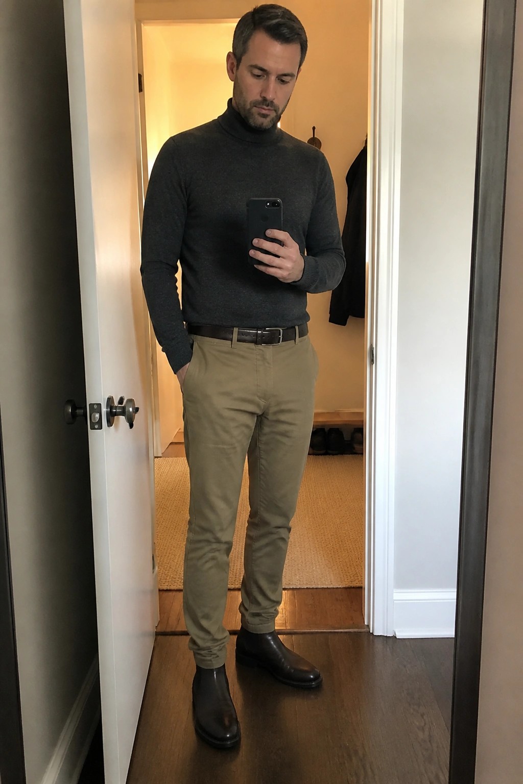 Man in gray turtleneck, khaki pants, and brown boots taking mirror selfie
