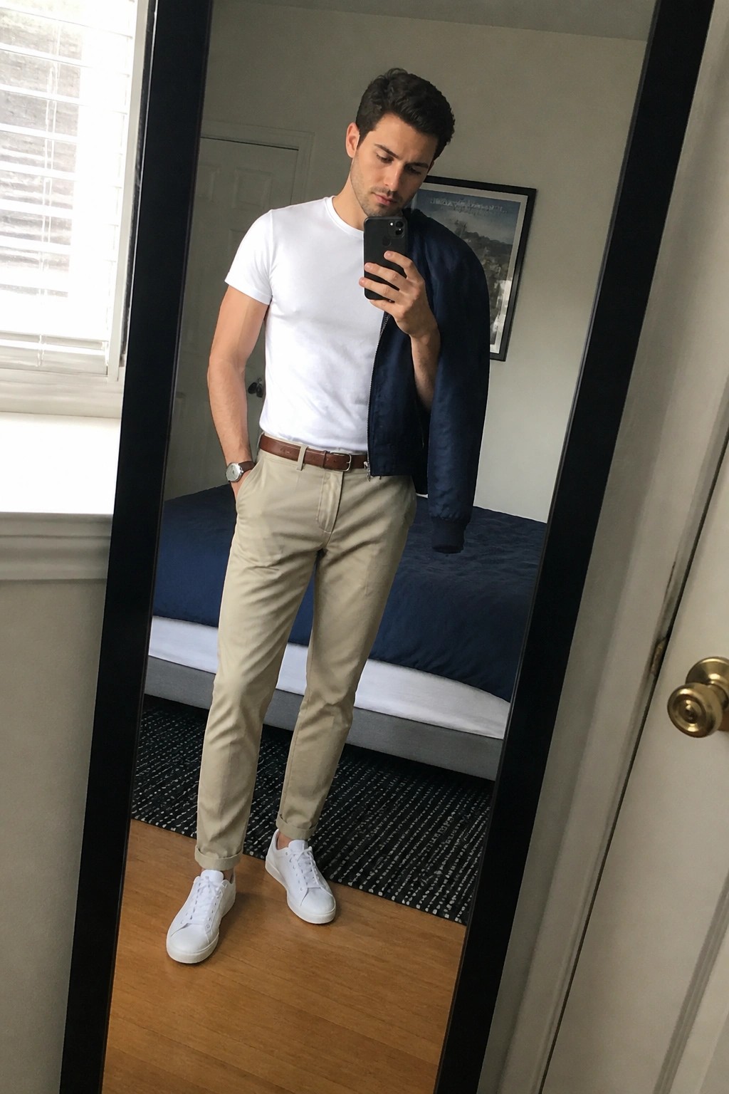 Man in mirror selfie wearing white crew neck t-shirt, beige khaki pants with brown belt, white sneakers, and navy bomber jacket draped over one shoulder.