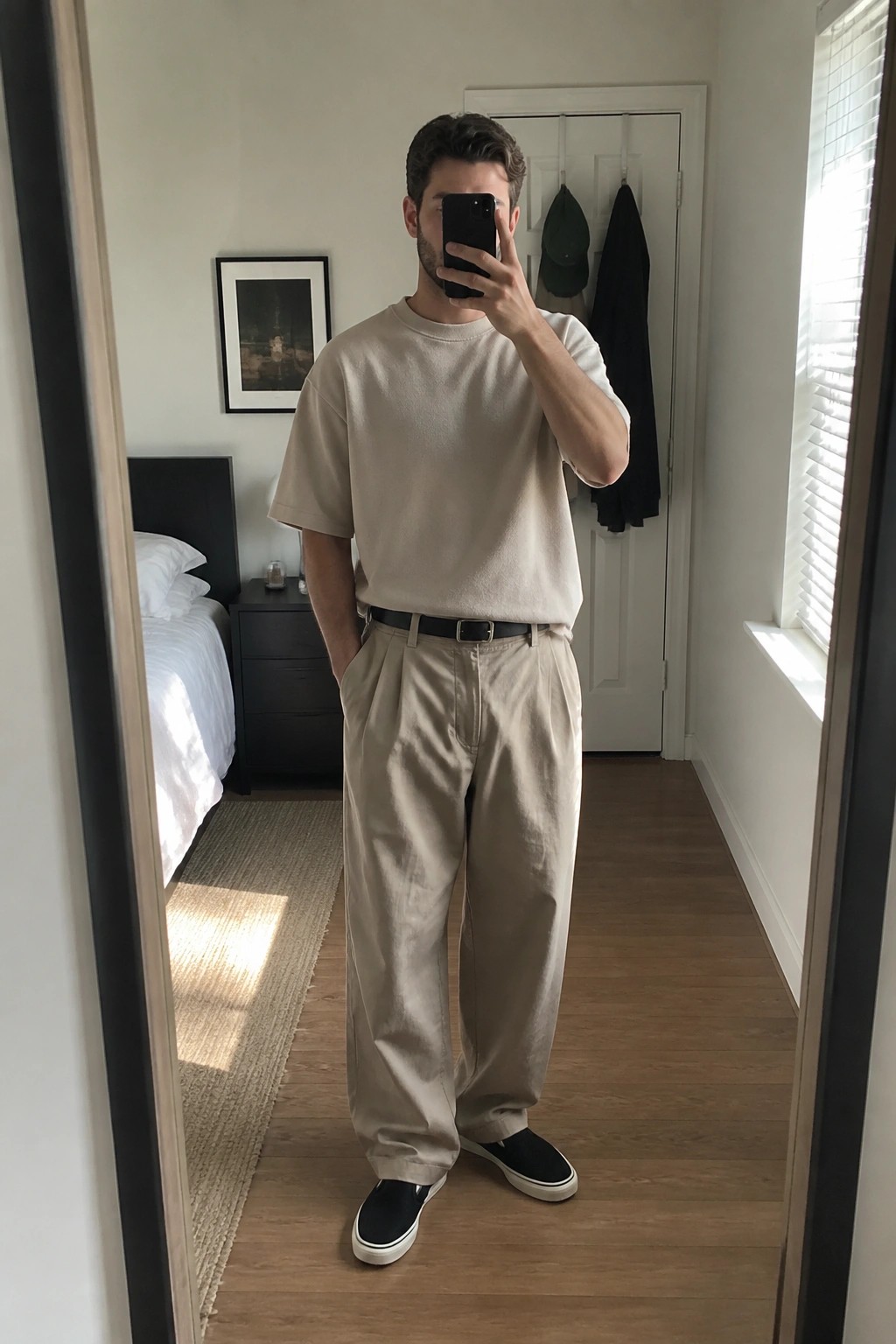 Man in beige t-shirt and pleated pants taking mirror selfie in bedroom.