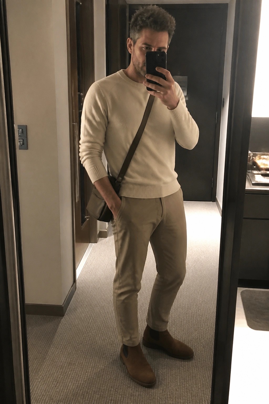 Man wearing a cream crewneck sweater tucked into slim khaki pants with brown suede Chelsea boots and a brown leather crossbody bag in a minimalist neutral palette.