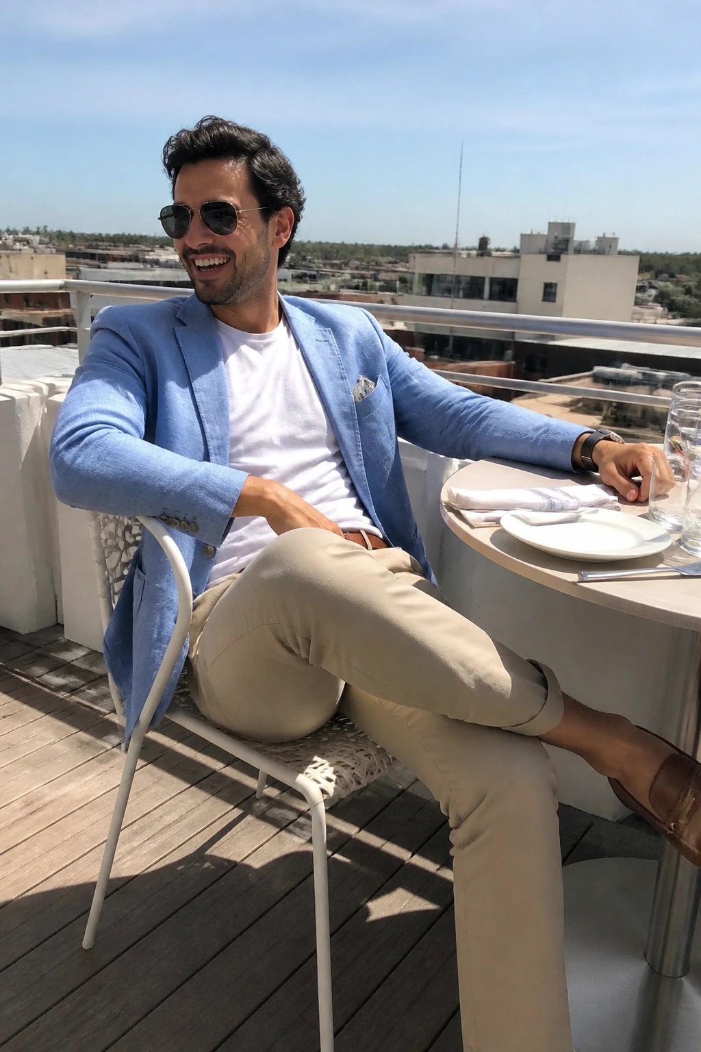 Man in blue blazer and beige pants smiling at sunny rooftop table.