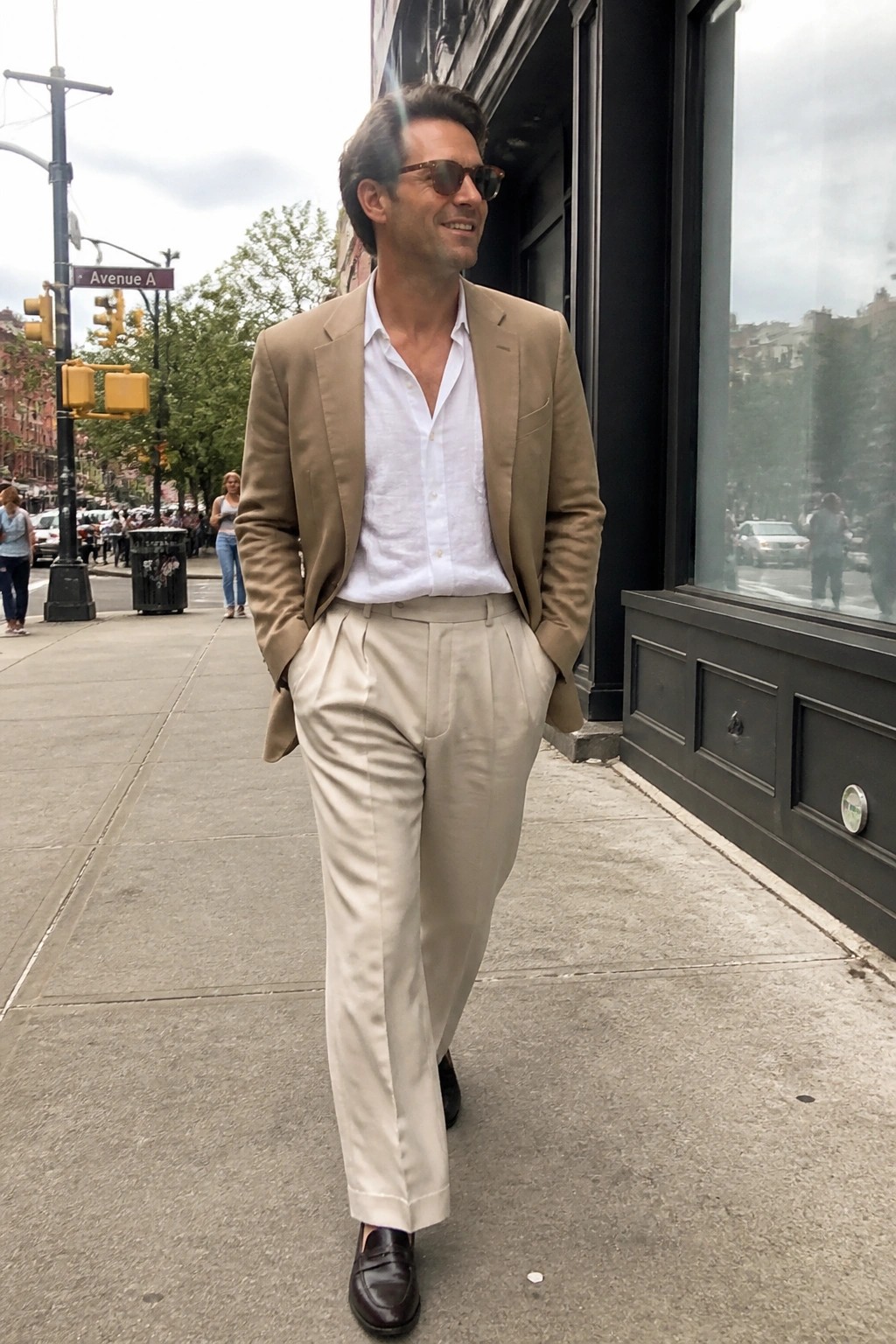 Stylish man in tan blazer and pleated trousers walking on city sidewalk.