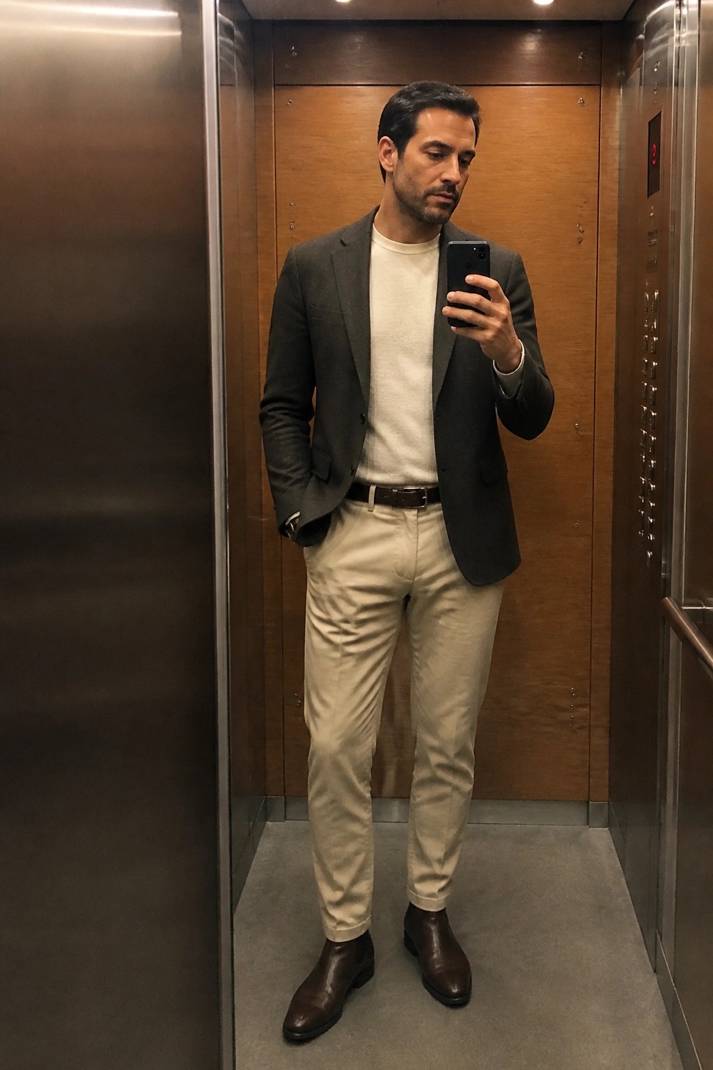 Man in gray blazer and beige pants taking mirror selfie in elevator.