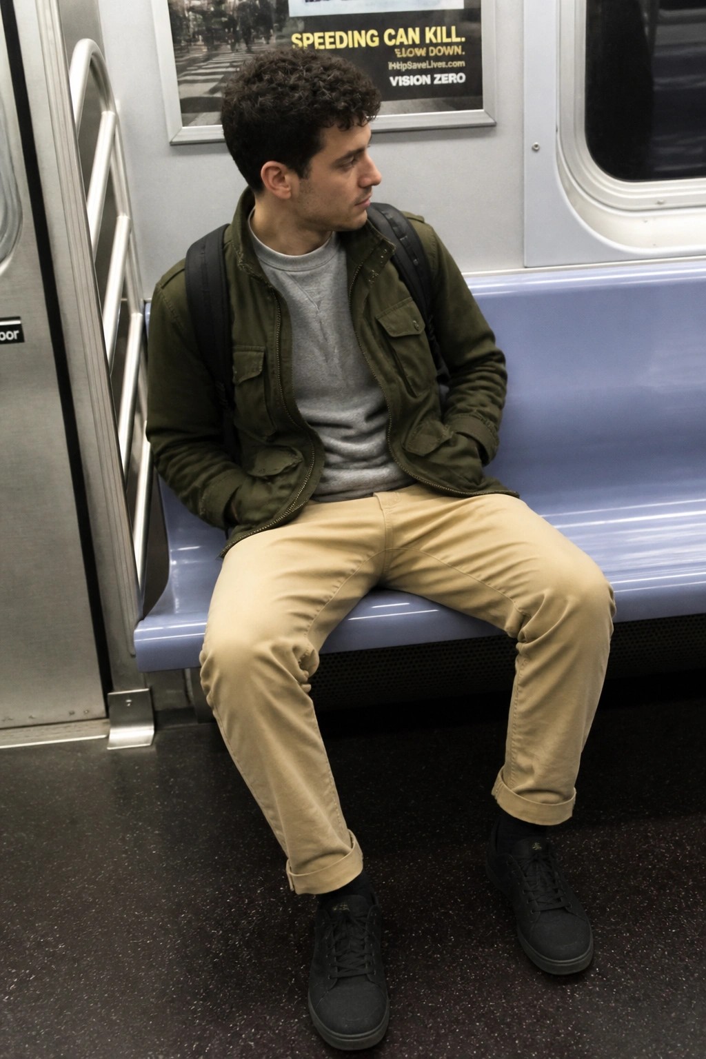 Young man in green jacket and khaki pants sits on blue subway bench.