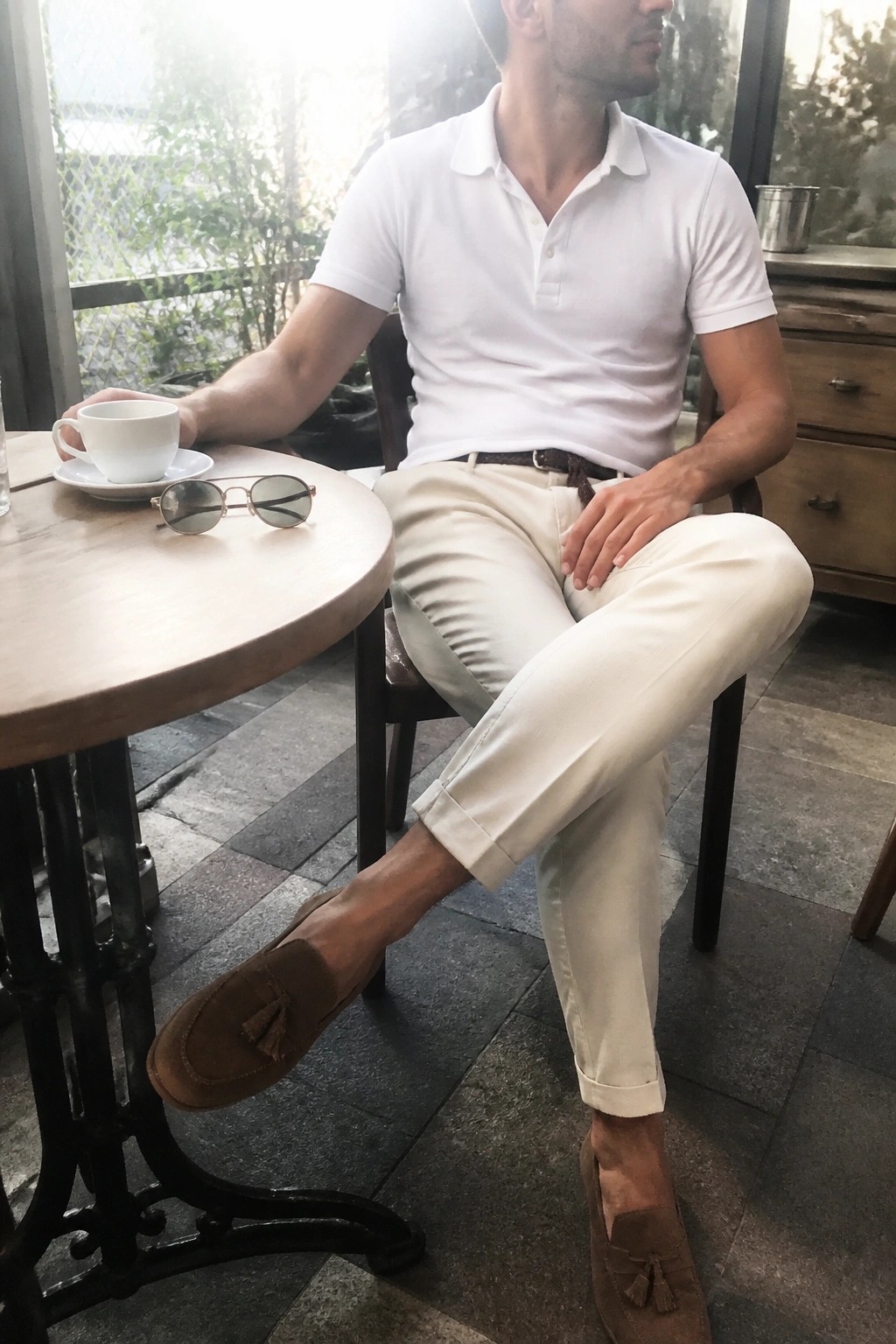 Man in white polo shirt and beige pants seated at wooden cafe table with sunglasses.