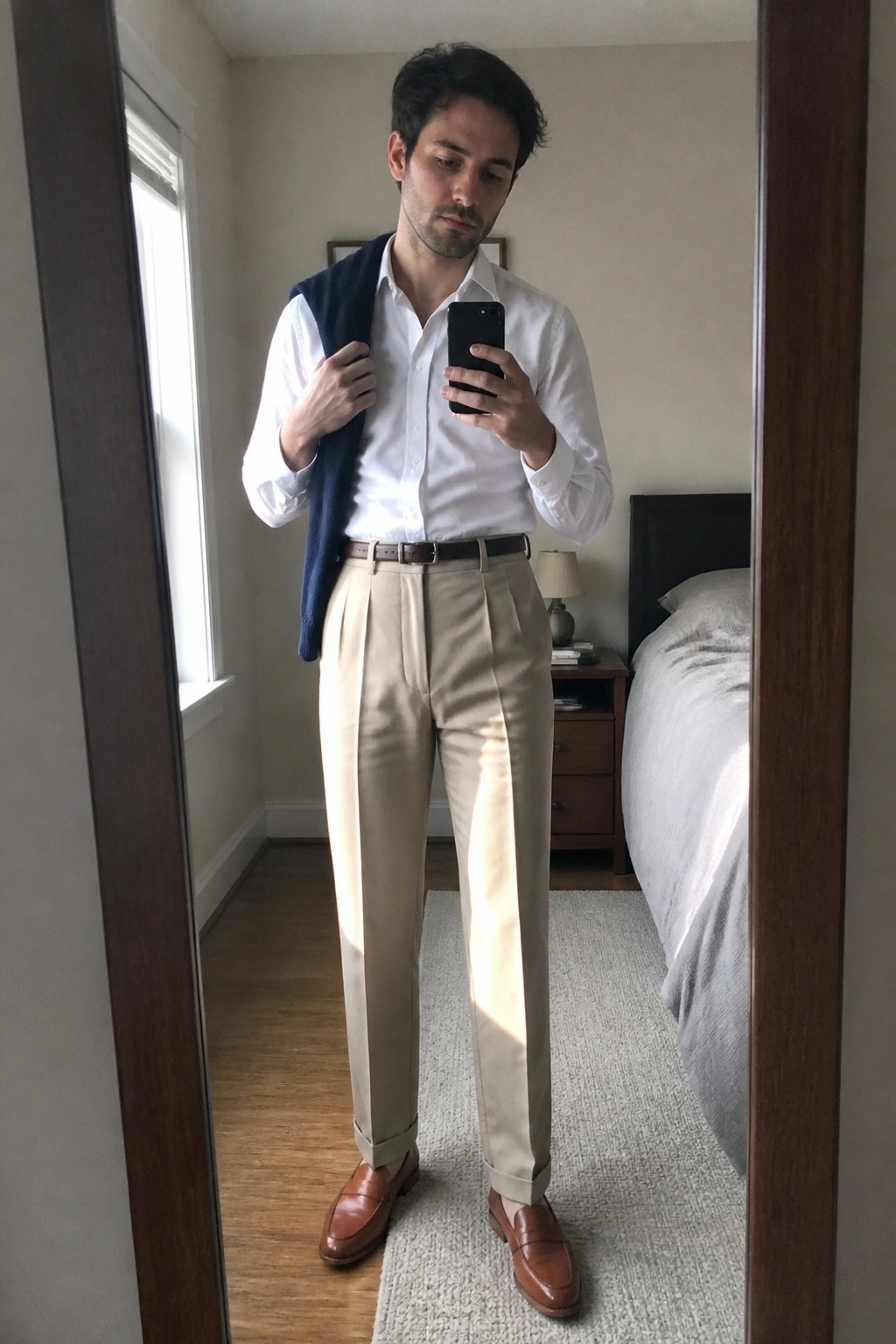 Man in white shirt and beige pleated pants holds phone with navy vest over shoulder