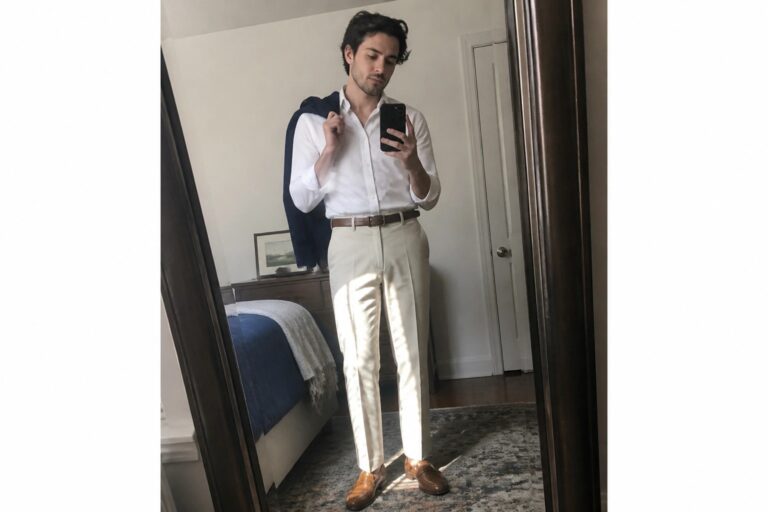 Man in white shirt and beige pleated pants holds phone with navy vest over shoulder