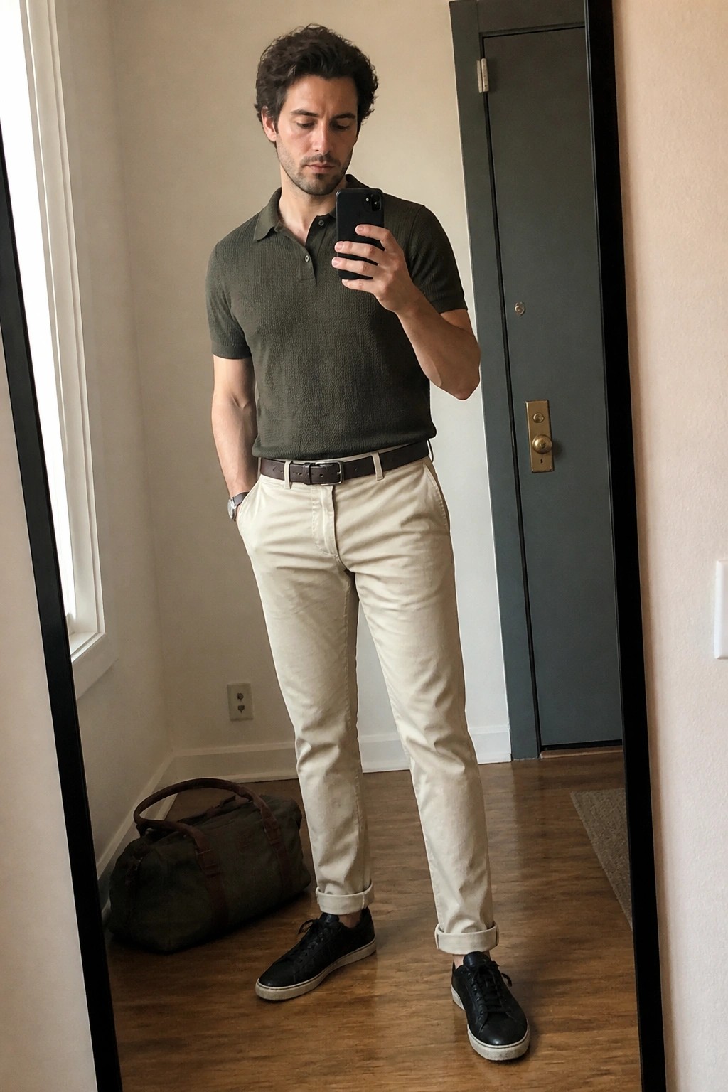 Man in olive polo and beige pants taking mirror selfie with phone
