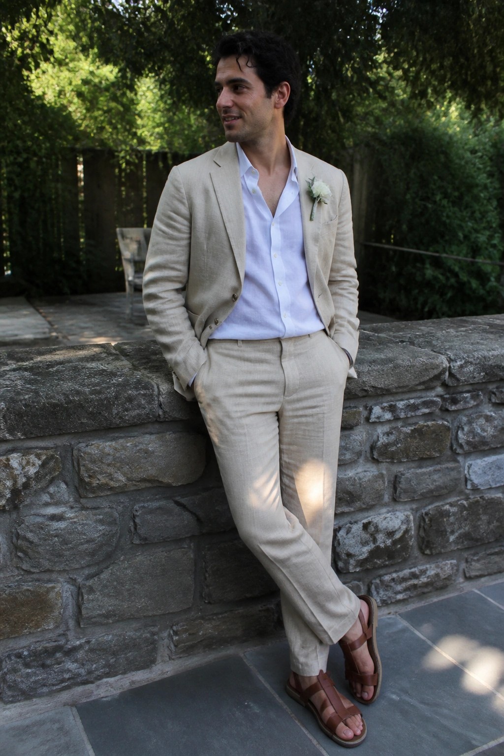 Man in beige linen suit with brown sandals leaning on stone garden wall