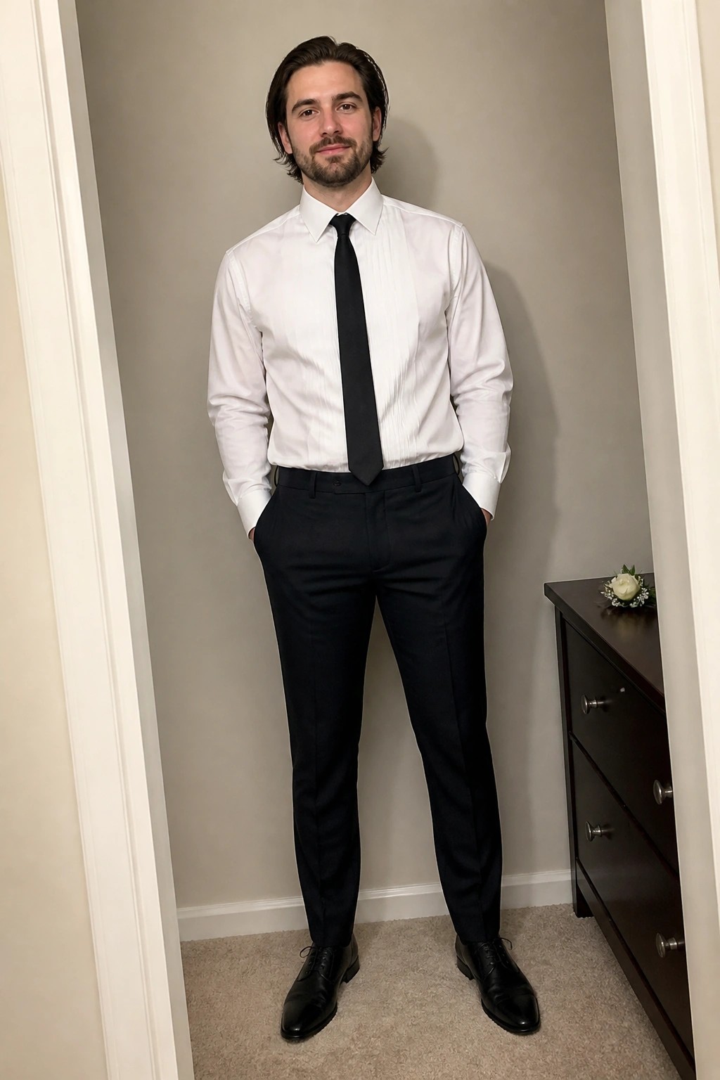 Man standing in black suit trousers, white pleated dress shirt, black tie, and black dress shoes with hands in pockets.