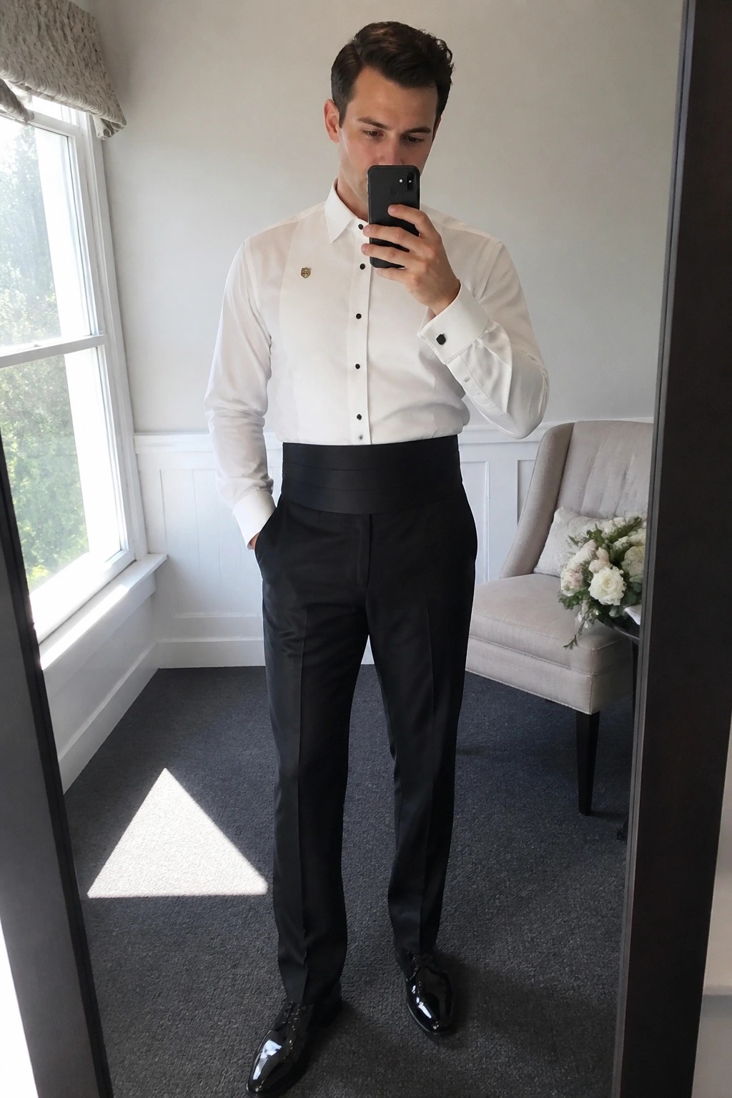 Man in white dress shirt with black cummerbund black trousers and patent shoes taking a mirror selfie in formal attire.