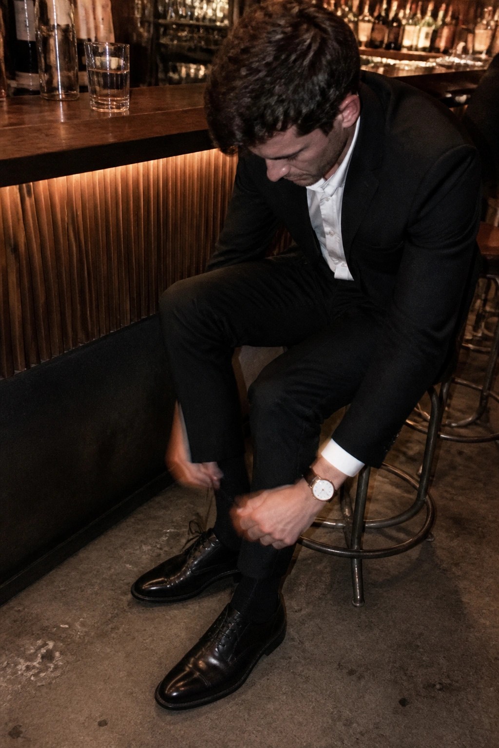 Man in black suit tying black dress shoe on stool at dimly lit bar