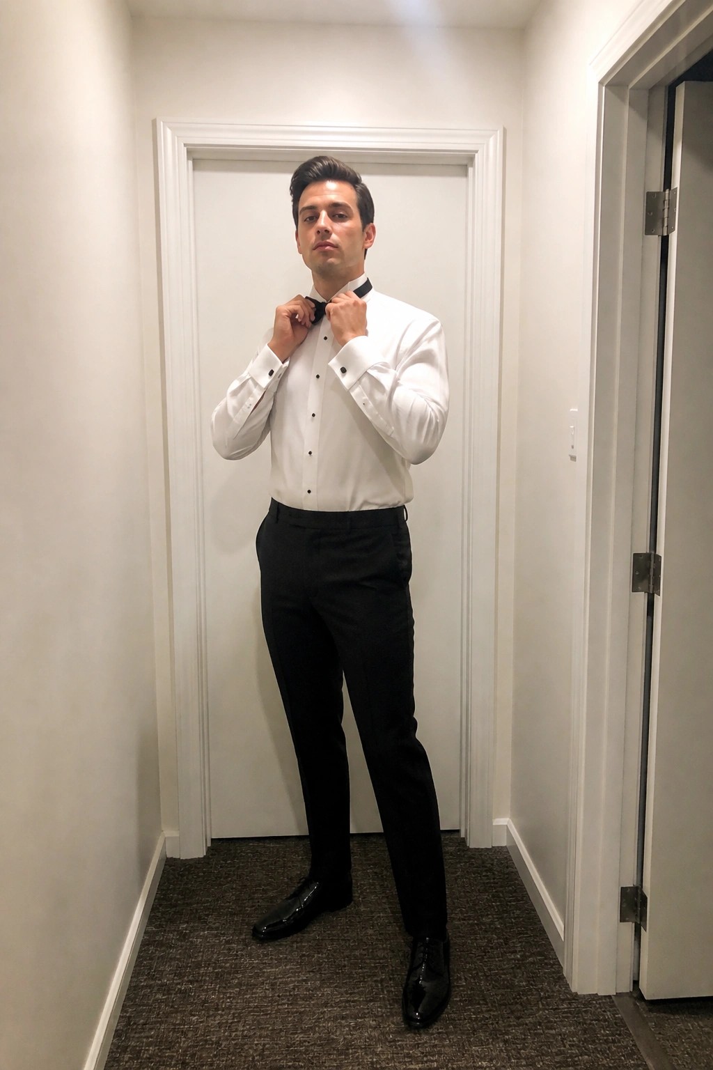 Man in white shirt and black bow tie adjusts collar in hallway