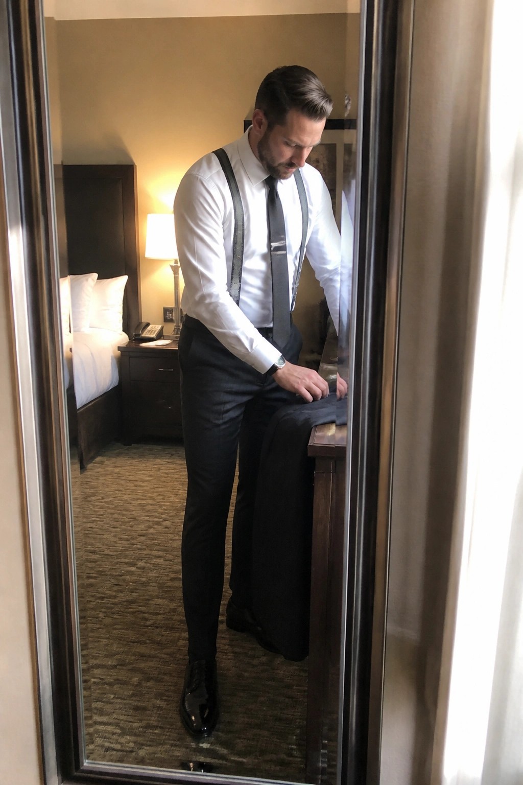 Man in white shirt, black tie, and suspenders adjusting clothes in hotel mirror