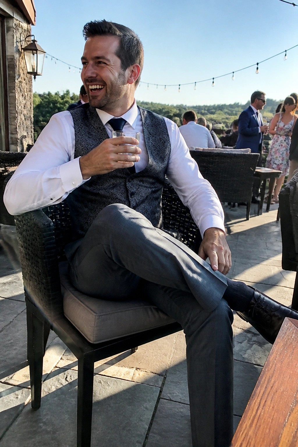 Man seated in gray trousers, patterned gray waistcoat, white dress shirt, black tie and black dress shoes while holding a glass at an outdoor event.