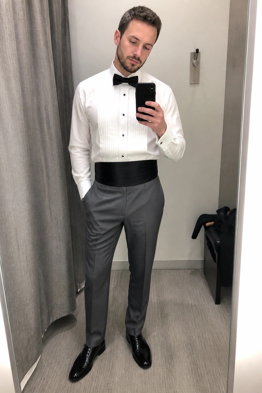 Man in white tuxedo shirt, black bow tie, cummerbund, and gray trousers taking selfie.