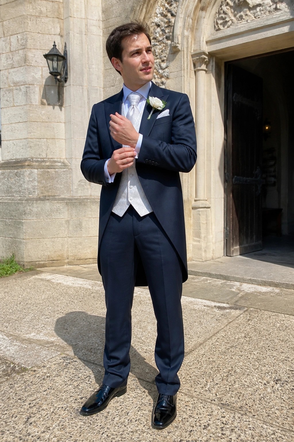 Man in navy tailcoat suit with silver patterned waistcoat and tie plus white boutonniere and black patent shoes standing on stone steps
