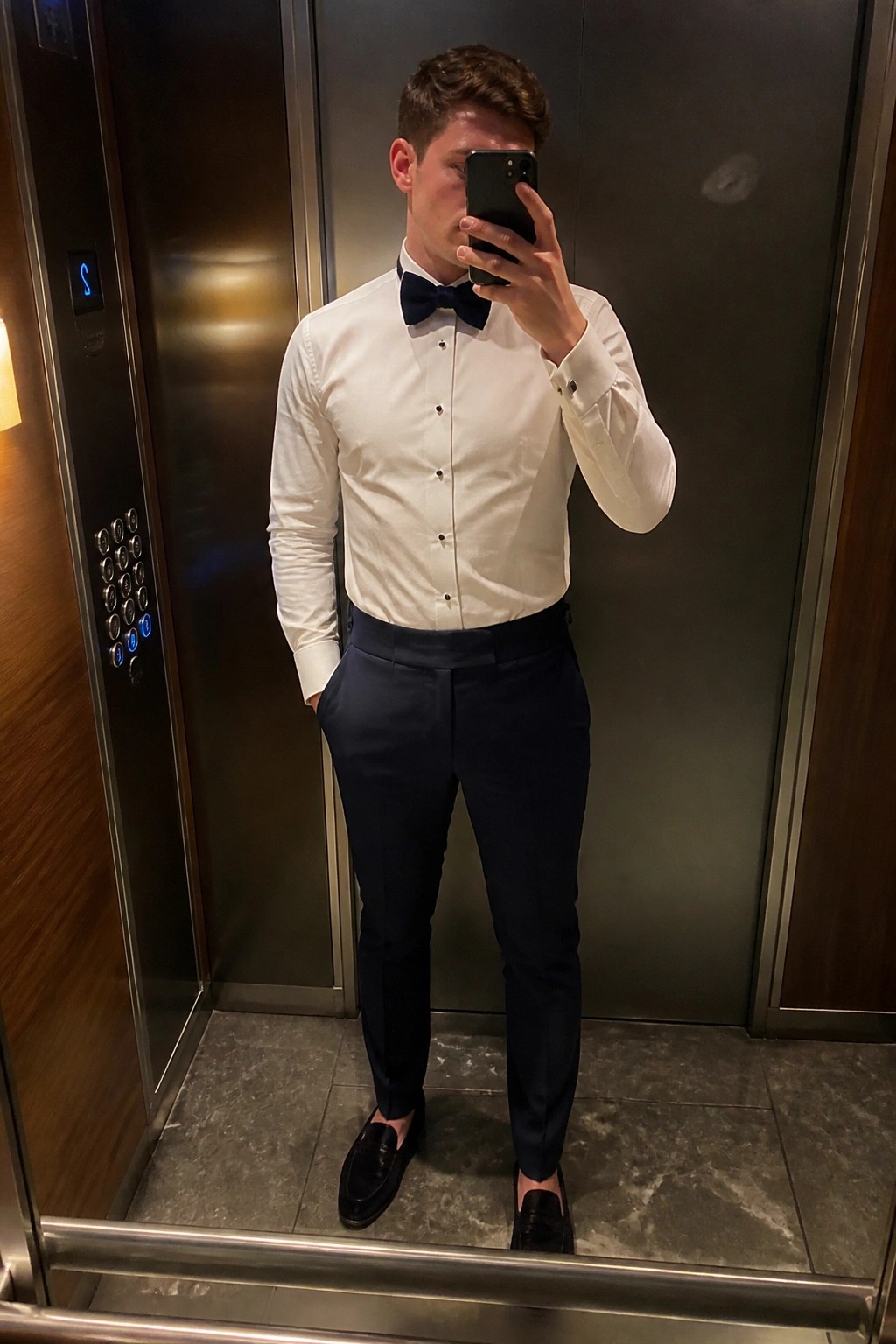 Man in white dress shirt black bow tie and navy trousers taking mirror selfie in elevator wearing black loafers