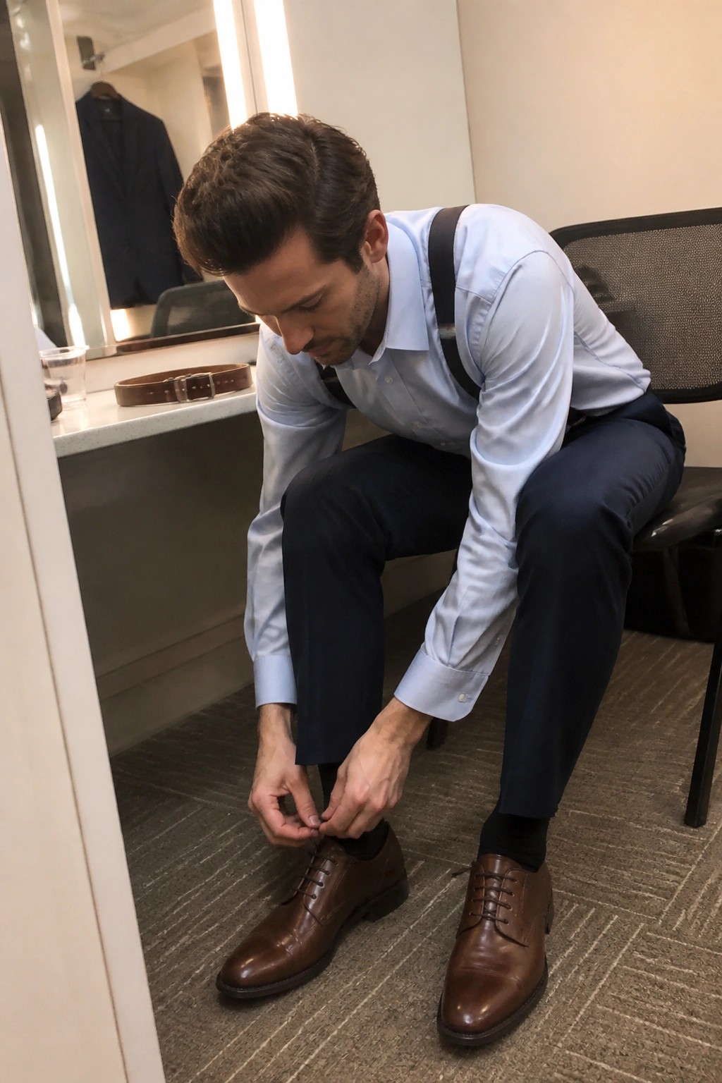 Man in light blue button down shirt navy trousers black suspenders and brown oxford shoes adjusting laces while seated in a dressing room.