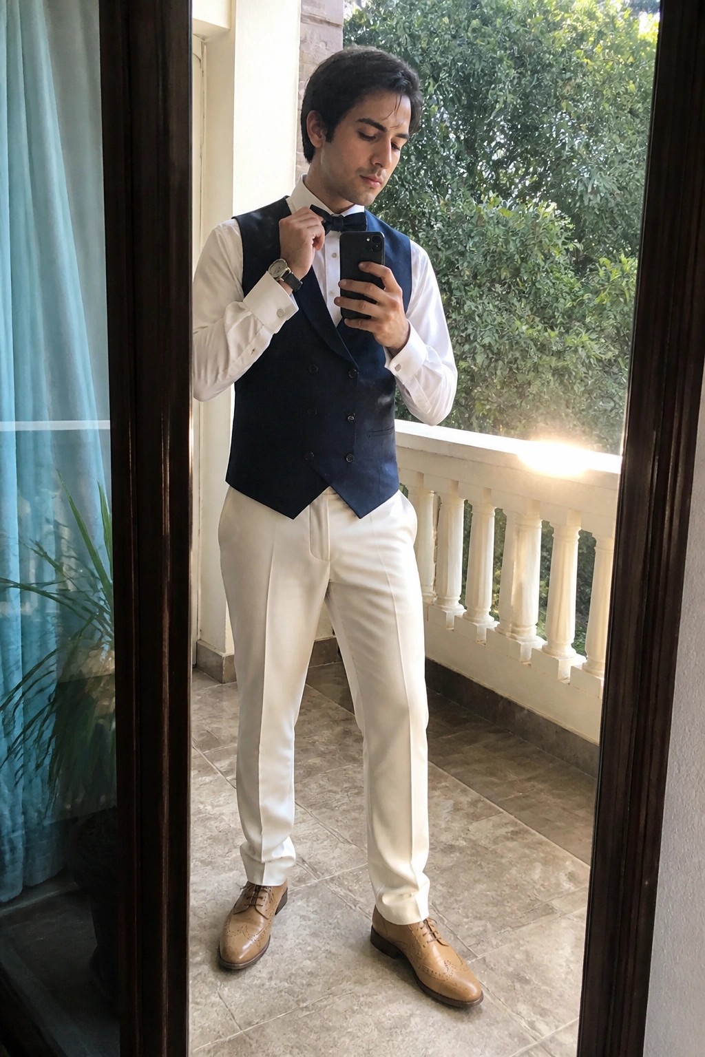 Man in navy vest and bow tie adjusts tie while viewing smartphone on balcony