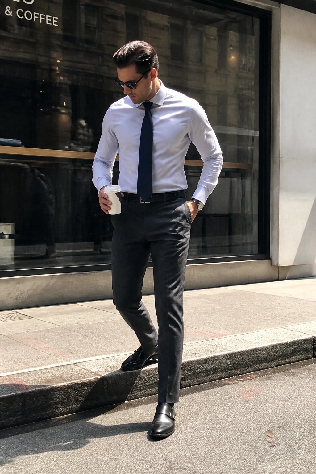 Man wearing light blue dress shirt, navy tie, dark grey formal pants, black belt, and black double monk strap shoes while holding a coffee cup on a city sidewalk.