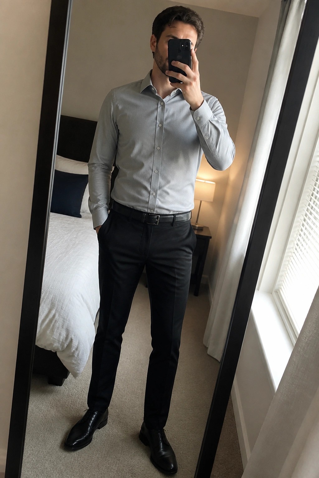 Man in light grey button down shirt tucked into black formal pants with black belt and black dress shoes taking a mirror selfie in a bedroom setting.
