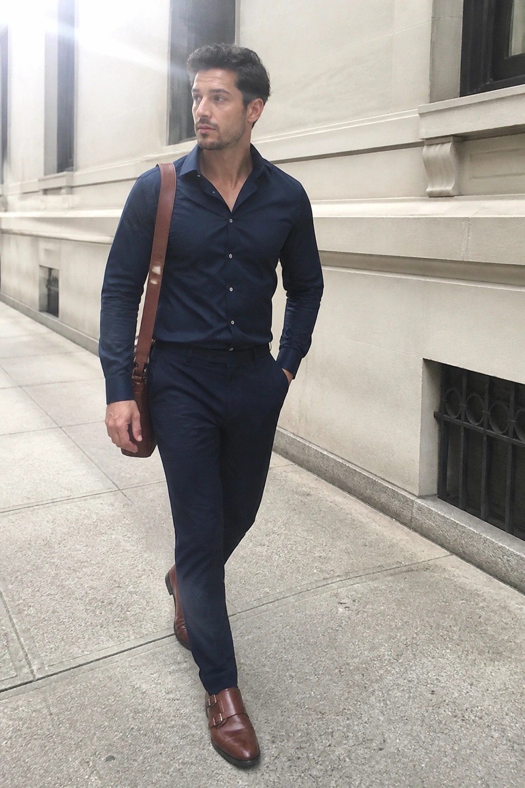 Man in navy shirt and pants with brown leather bag walking on sidewalk