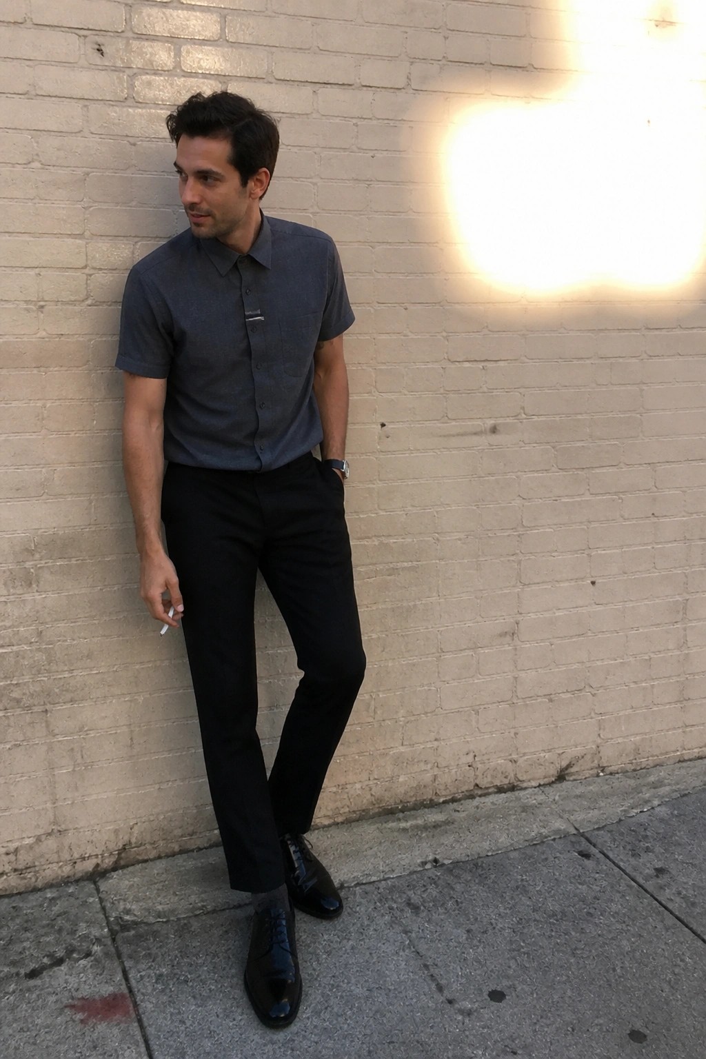 Man in gray short-sleeve shirt and black pants holds cigarette by brick wall