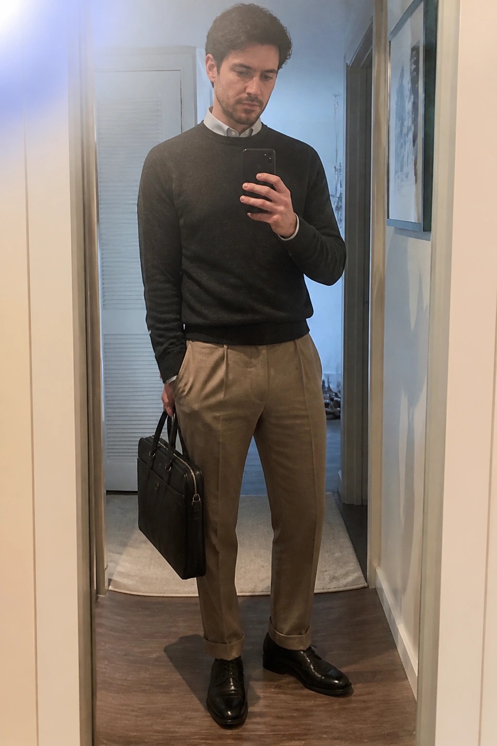 Man in gray sweater and tan pants holding phone and black briefcase in hallway