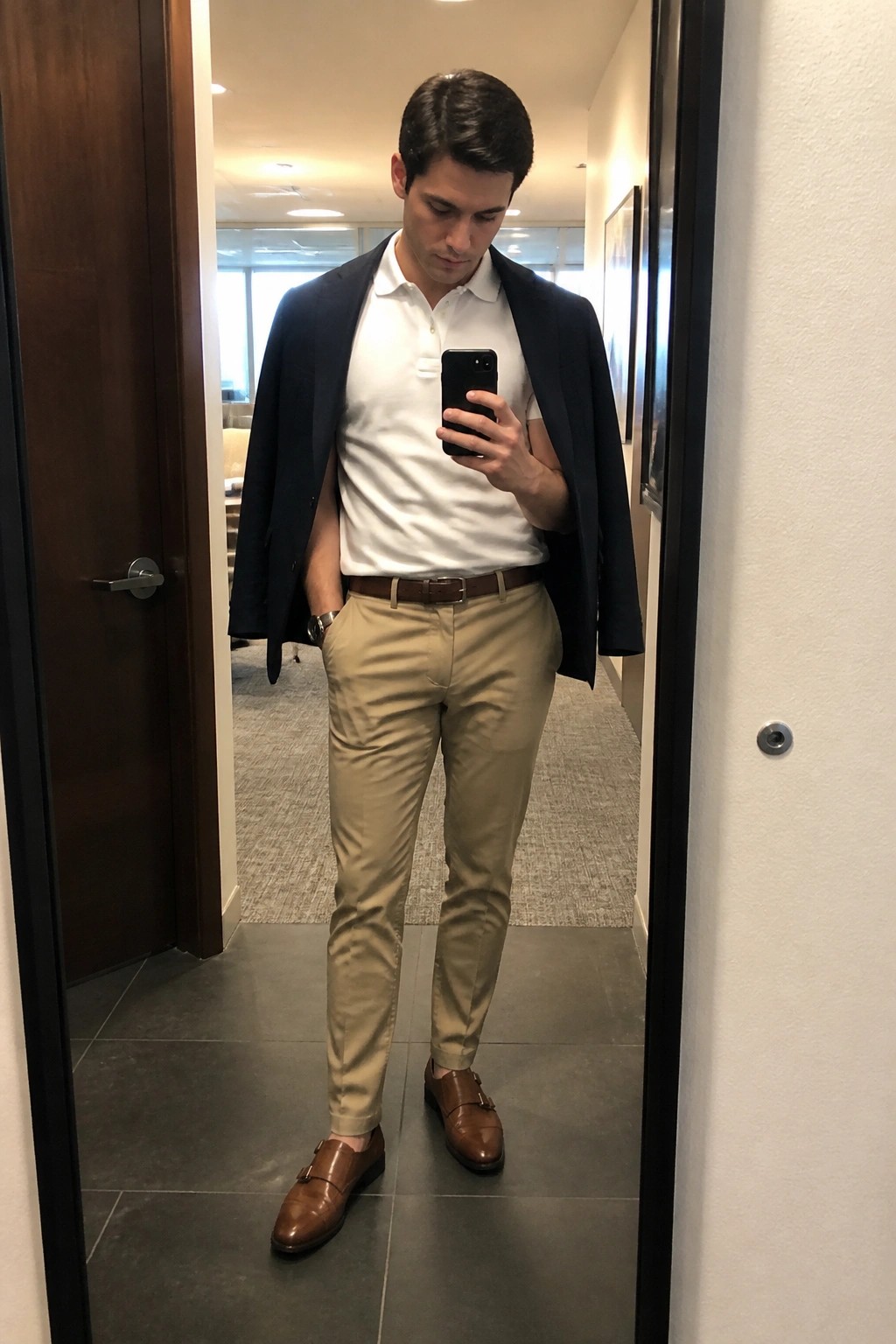 Man taking mirror selfie in office wearing white polo, khakis, and navy blazer over shoulders