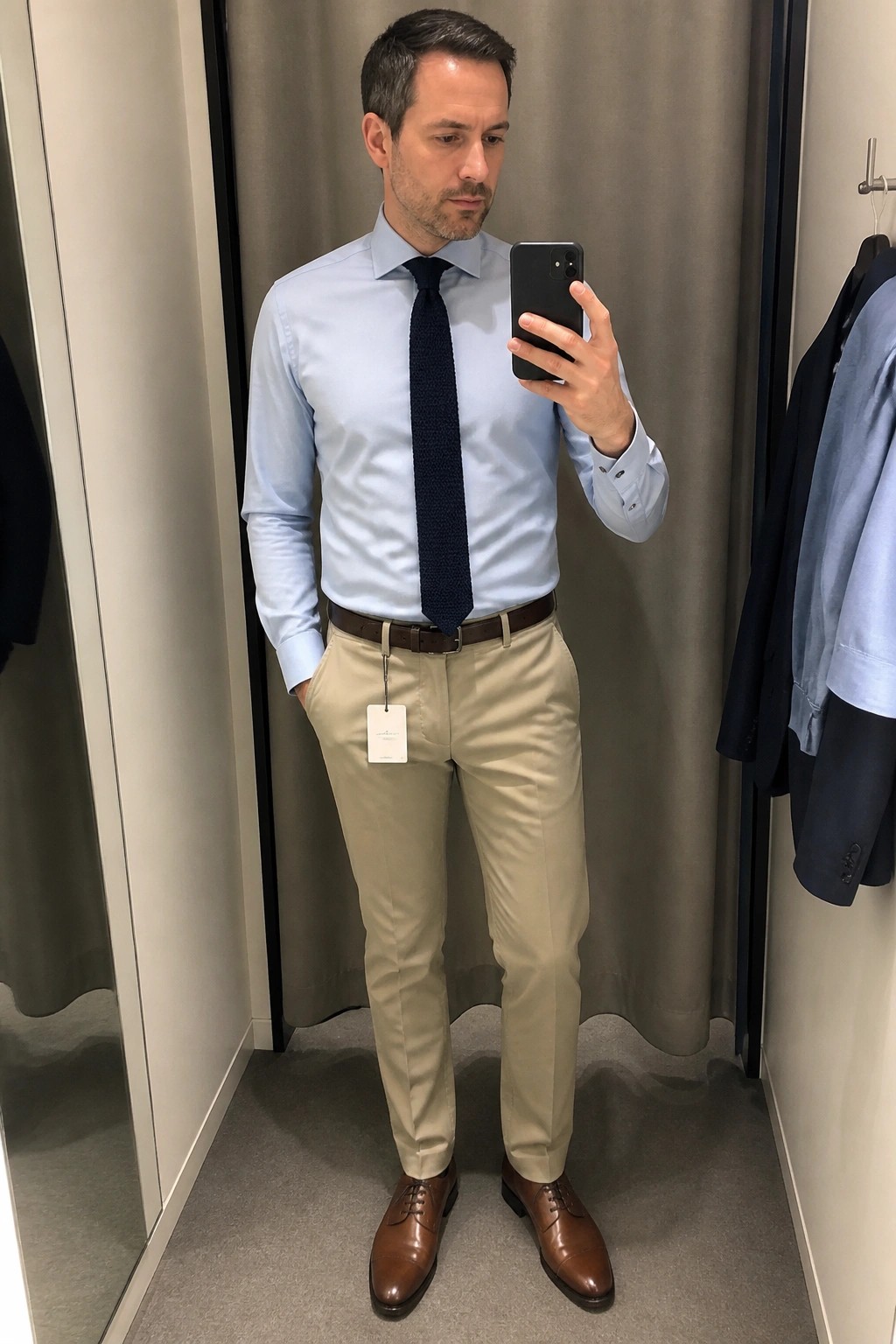 Man in light blue shirt and navy tie taking mirror selfie in fitting room