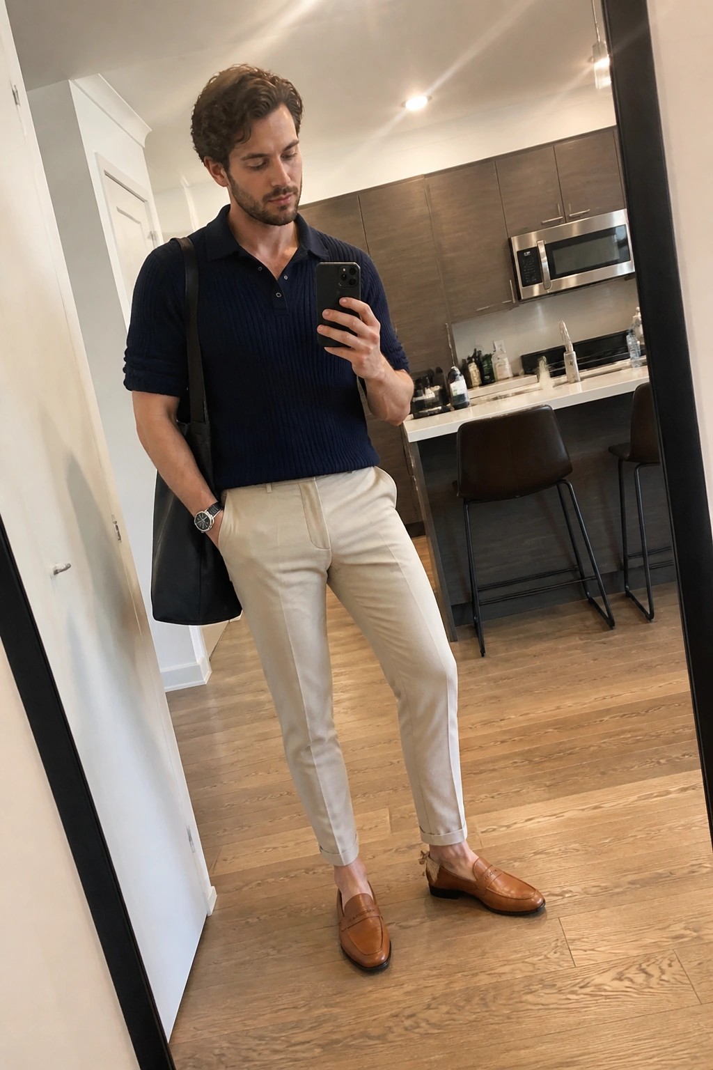 Man in navy polo and beige pants taking mirror selfie in modern kitchen