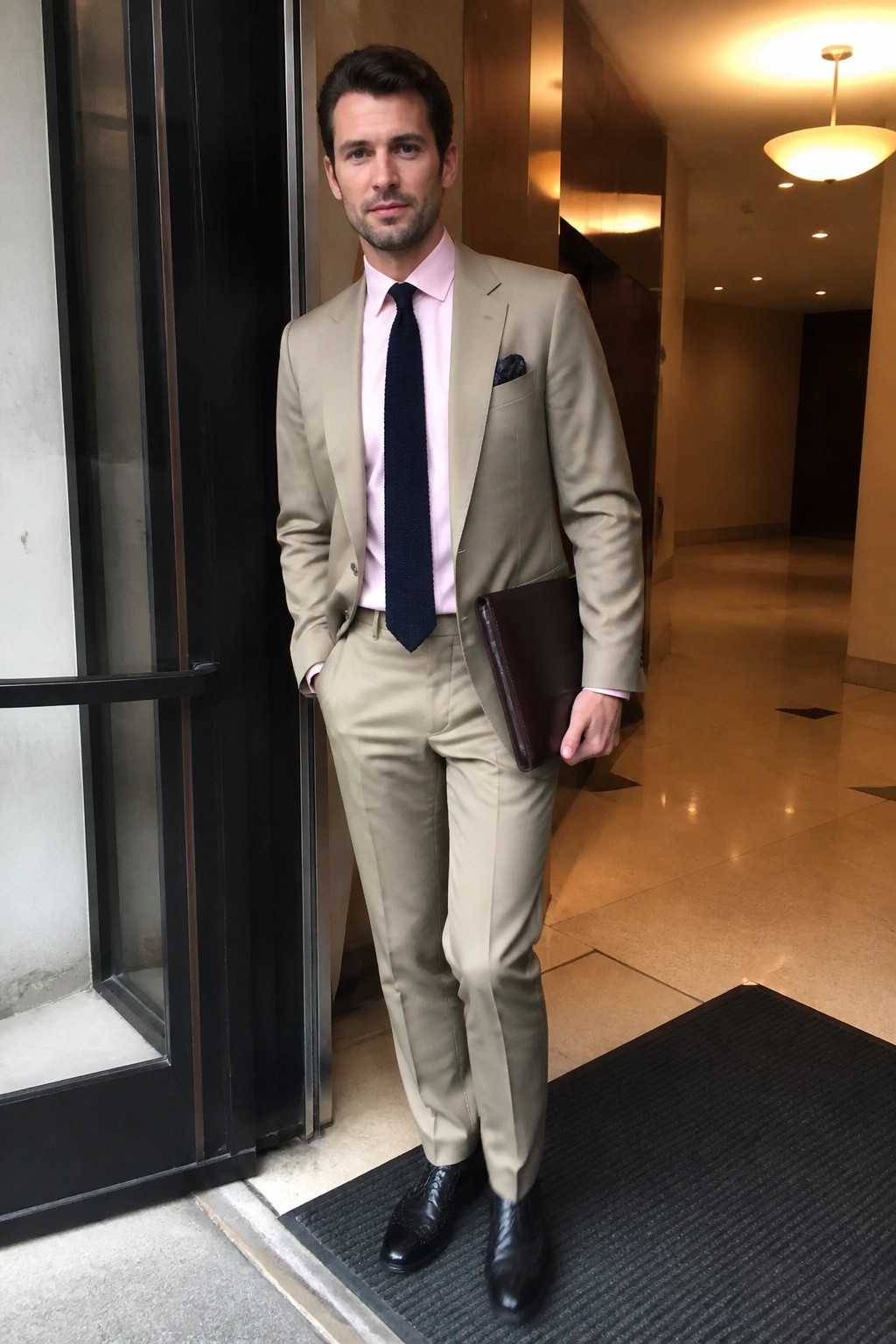 Man in beige suit, pink shirt and navy tie holding leather portfolio.