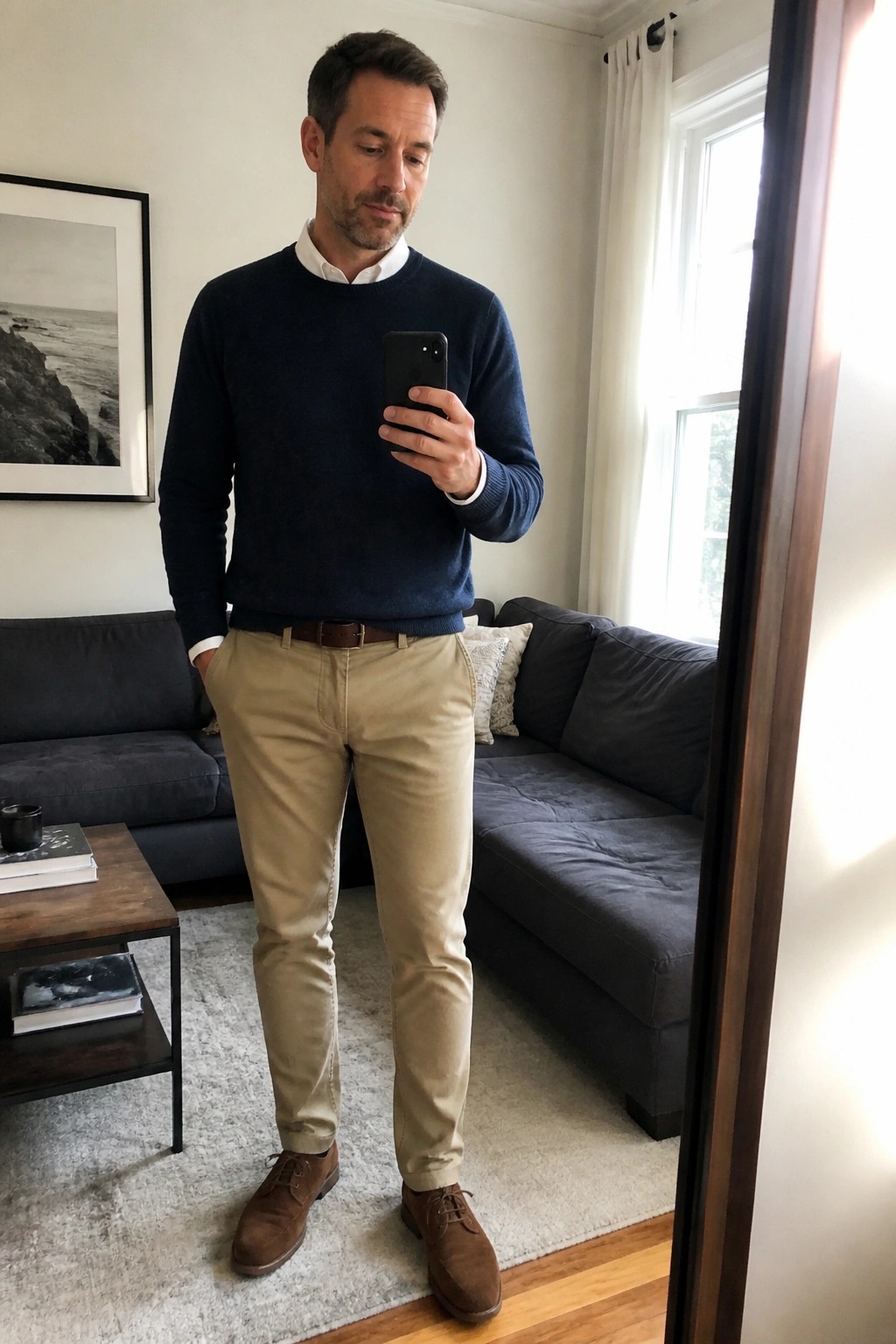 Man in navy sweater and khaki pants taking mirror selfie in living room