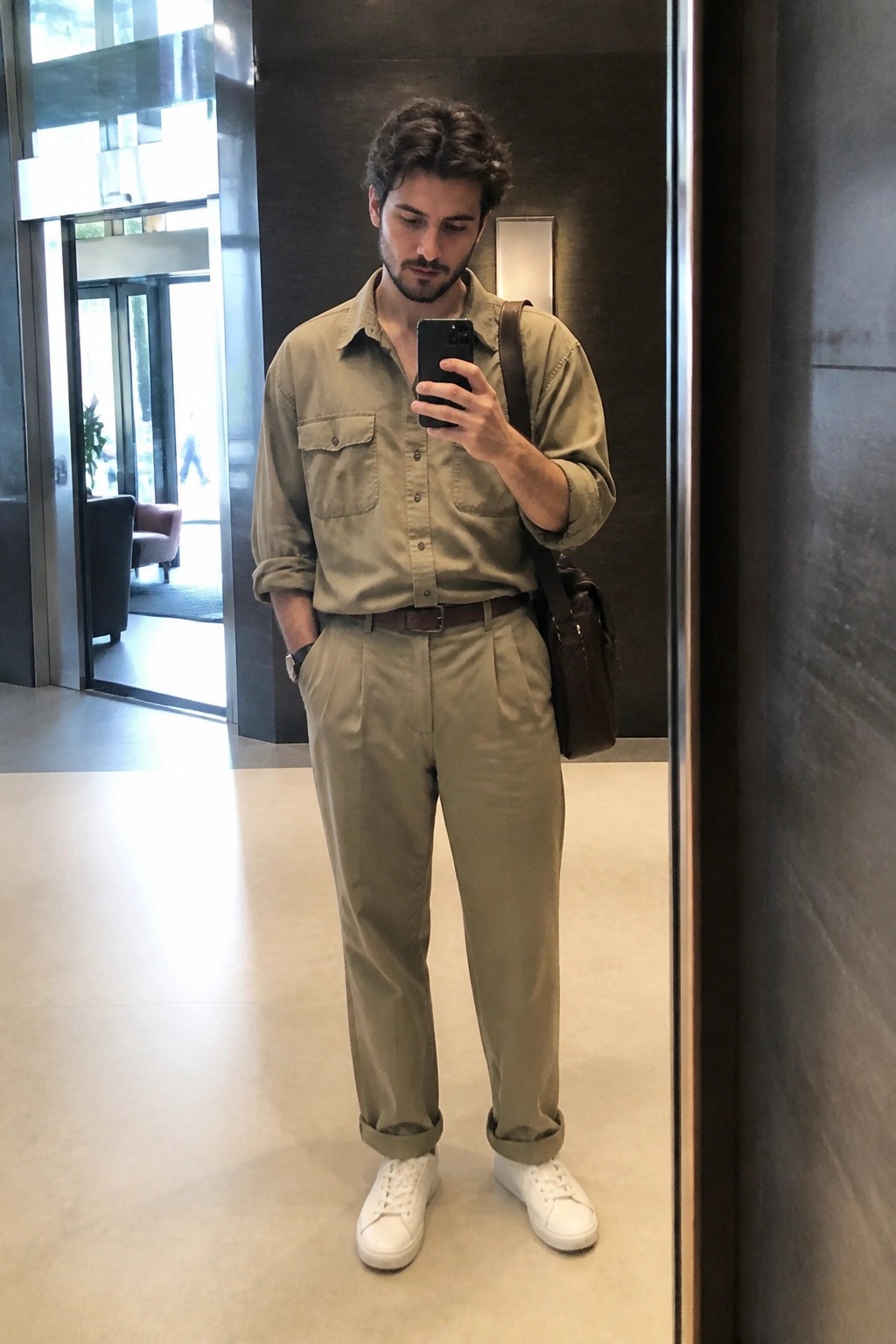 Man in khaki shirt and pleated pants taking mirror selfie with phone and bag.