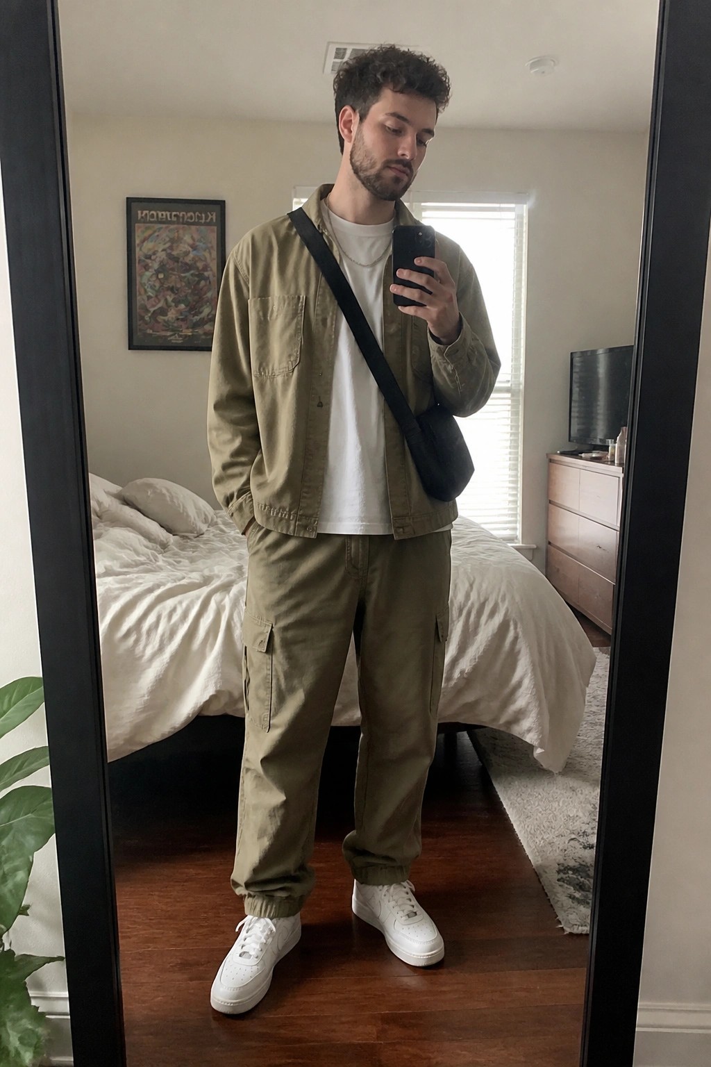 Man in oversized khaki utility jacket over white t-shirt with matching cargo pants, white sneakers, and black crossbody bag while holding a phone.