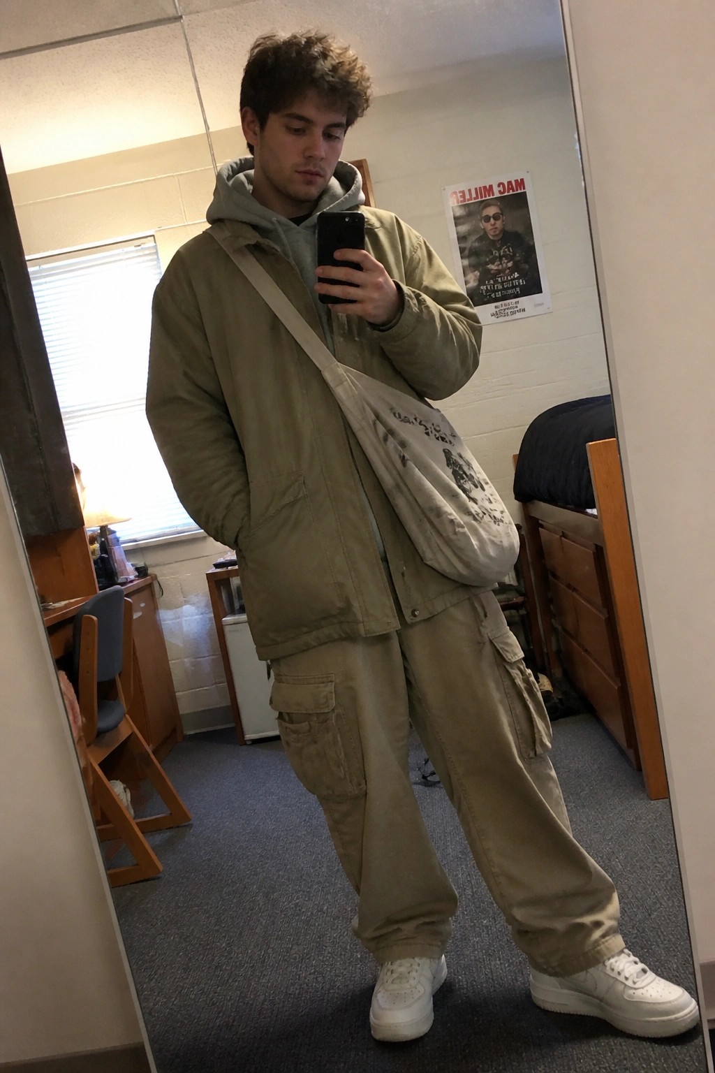 Young man in beige cargo outfit taking mirror selfie in dorm room