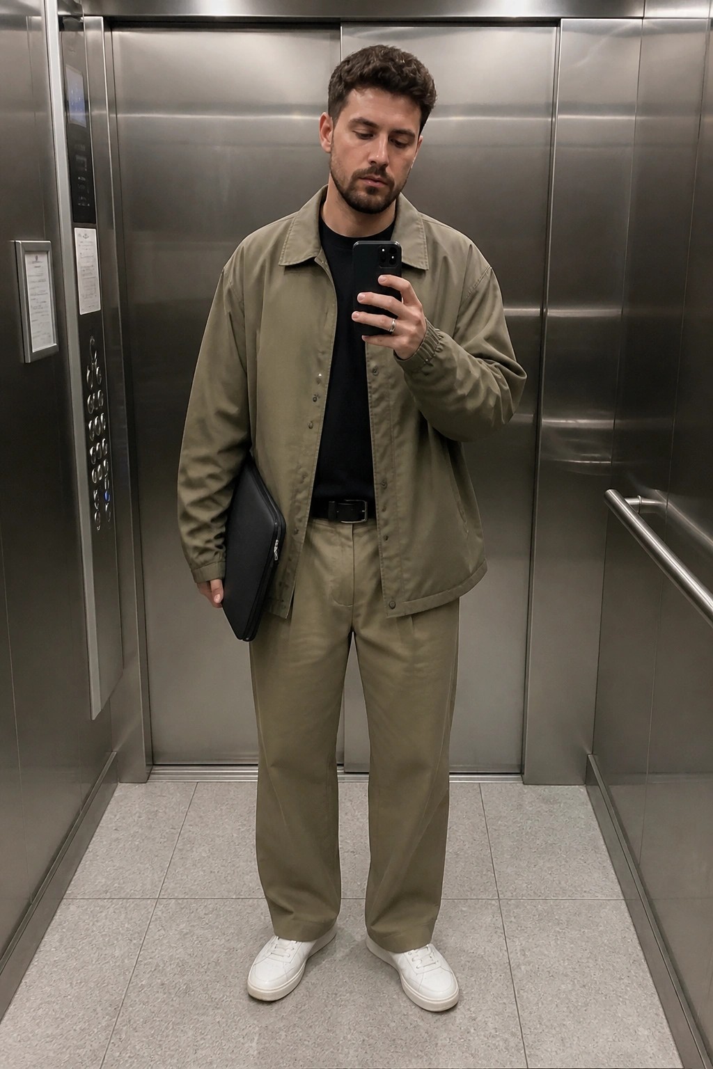 Man wearing an oversized khaki jacket over a black t-shirt with matching khaki trousers, black belt, white sneakers, and holding a black pouch while taking a mirror selfie in an elevator.