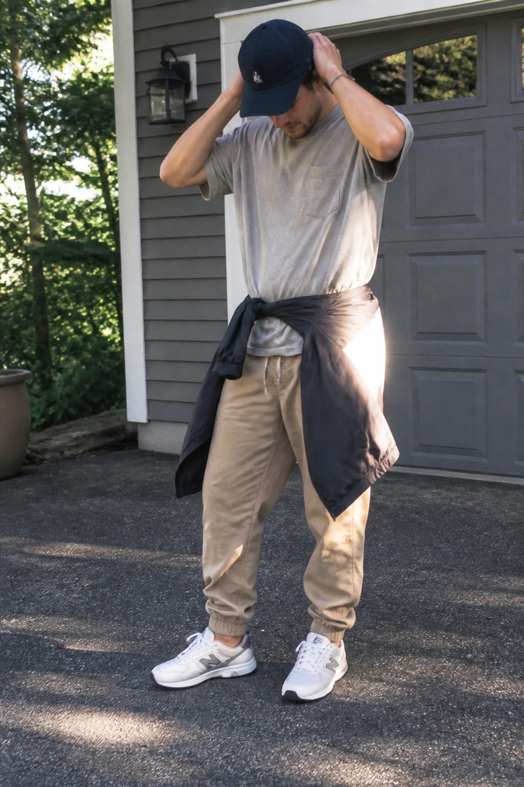 Man adjusting navy cap, wearing gray t-shirt, beige joggers, black shirt at waist.