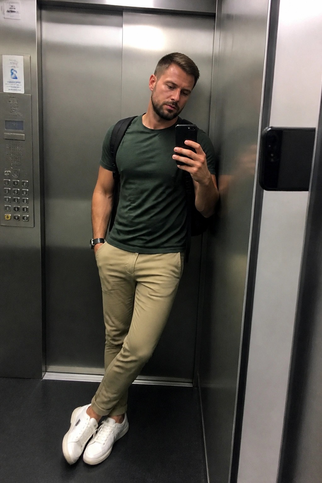 Man in green t-shirt and beige pants taking mirror selfie in elevator