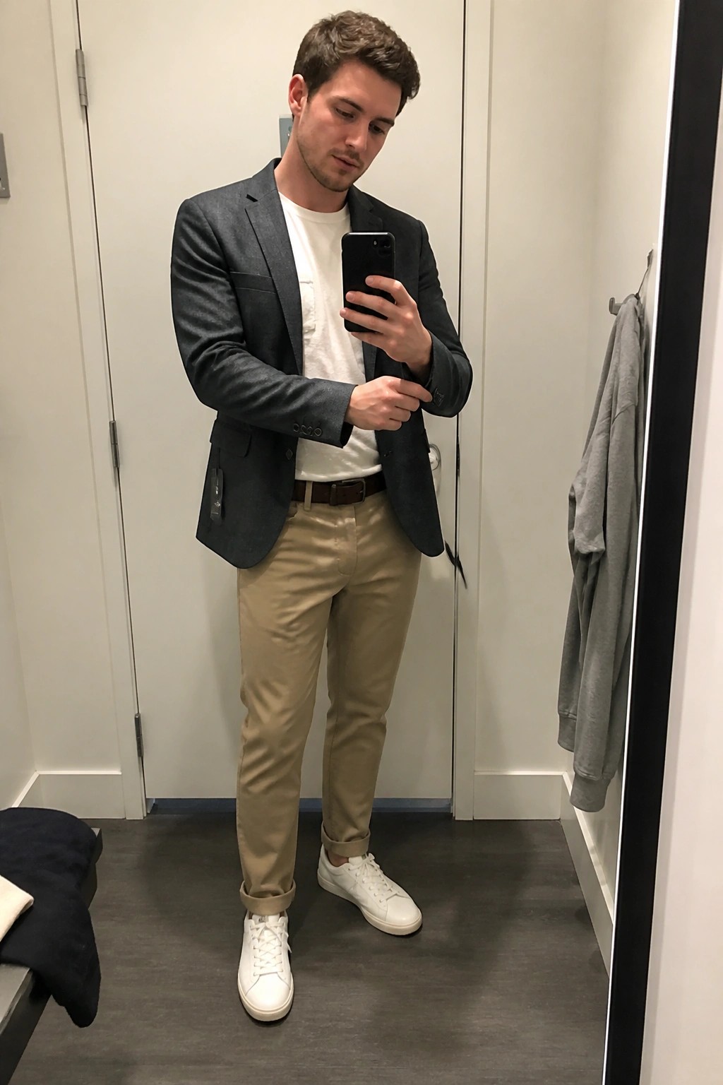 Man standing in a fitting room mirror selfie wearing a gray blazer over a white t-shirt with khaki pants, brown belt, and white sneakers while holding a phone.