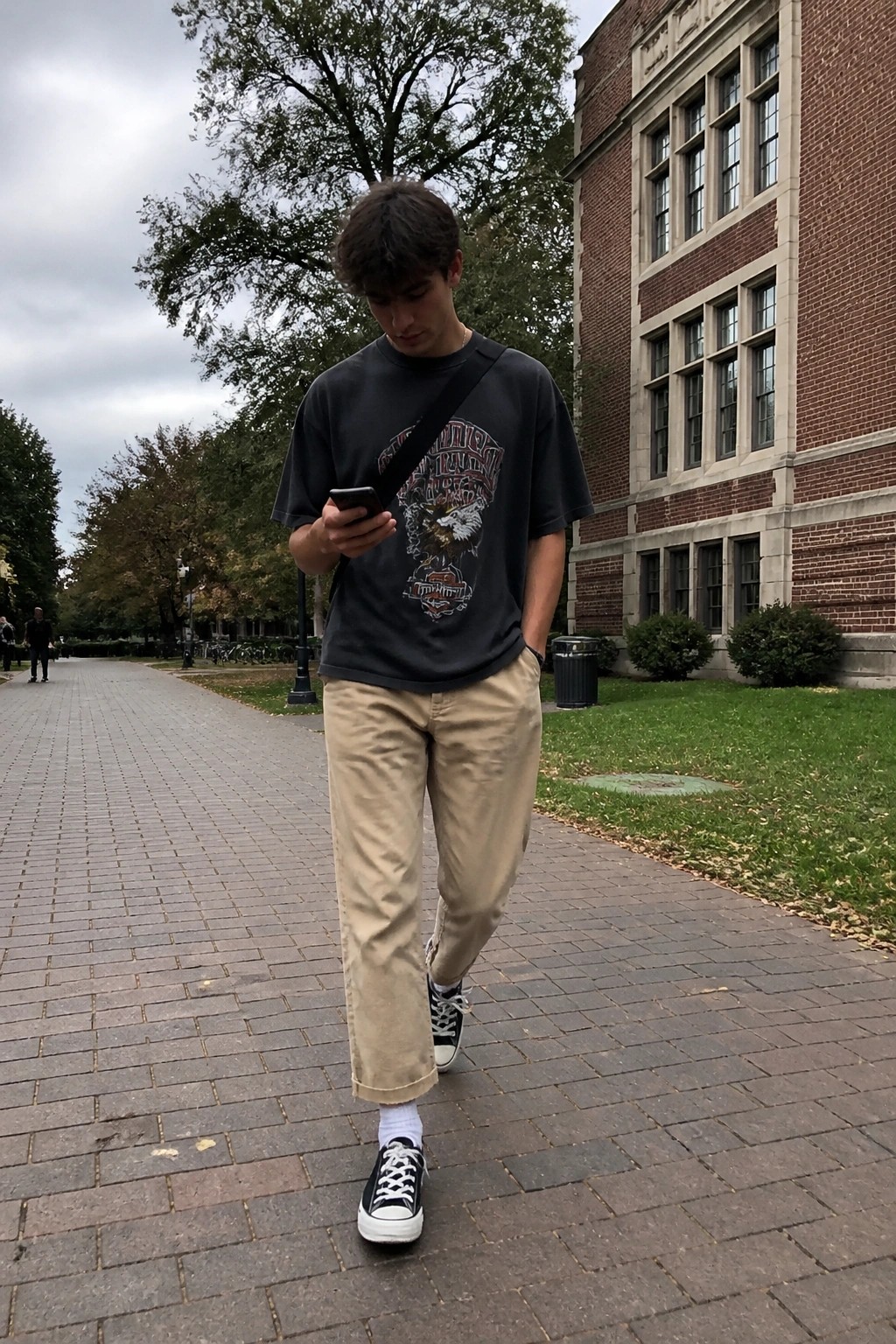 Young man wearing beige khaki pants cuffed at the ankle, a dark gray graphic print t-shirt, black and white low top Converse sneakers, and a black crossbody strap while walking on a brick path and checking his phone.