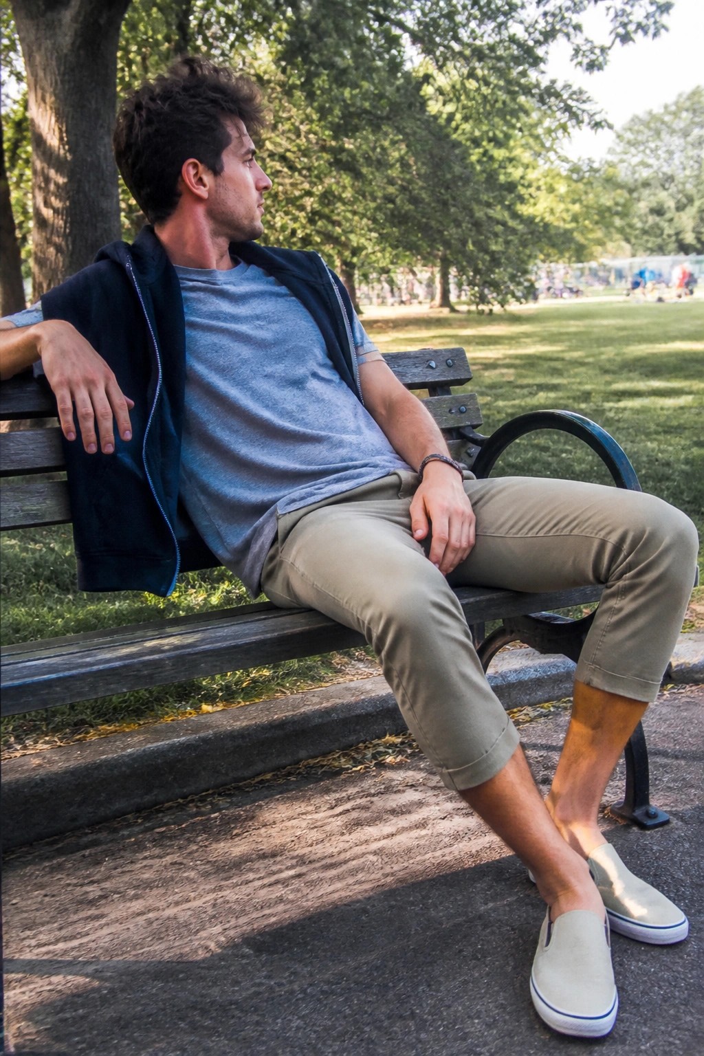 Man seated on park bench wearing khaki cropped pants, blue t-shirt, navy zip layer over shoulders, and light gray slip-on sneakers.