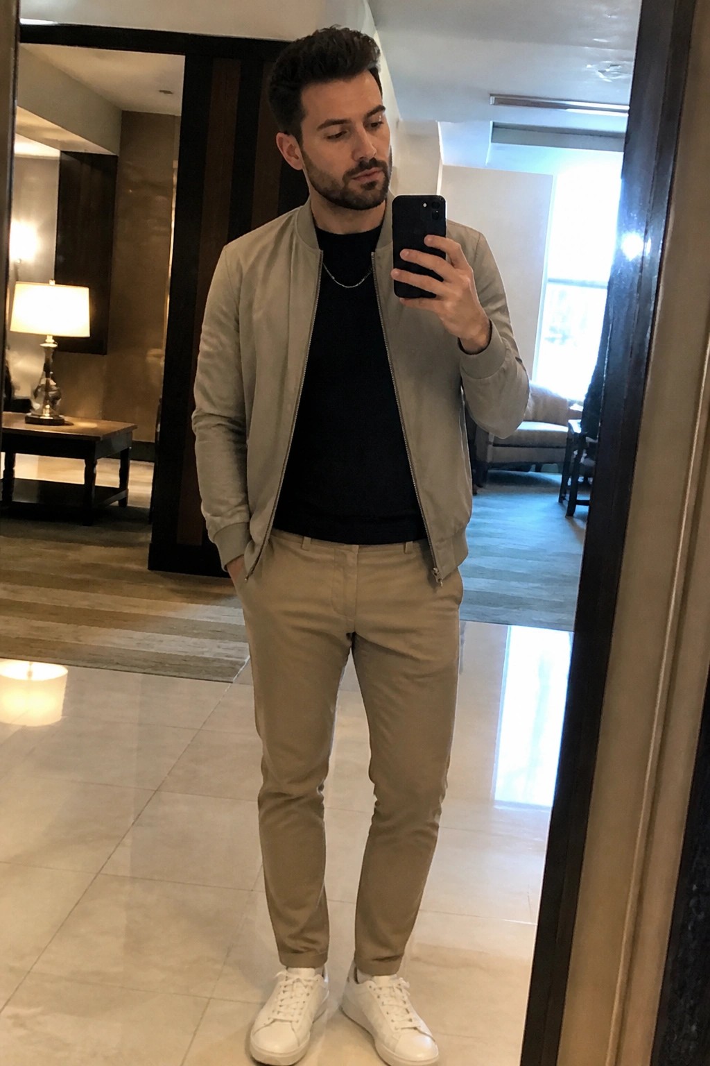 Bearded man in beige bomber jacket taking mirror selfie in hotel lobby