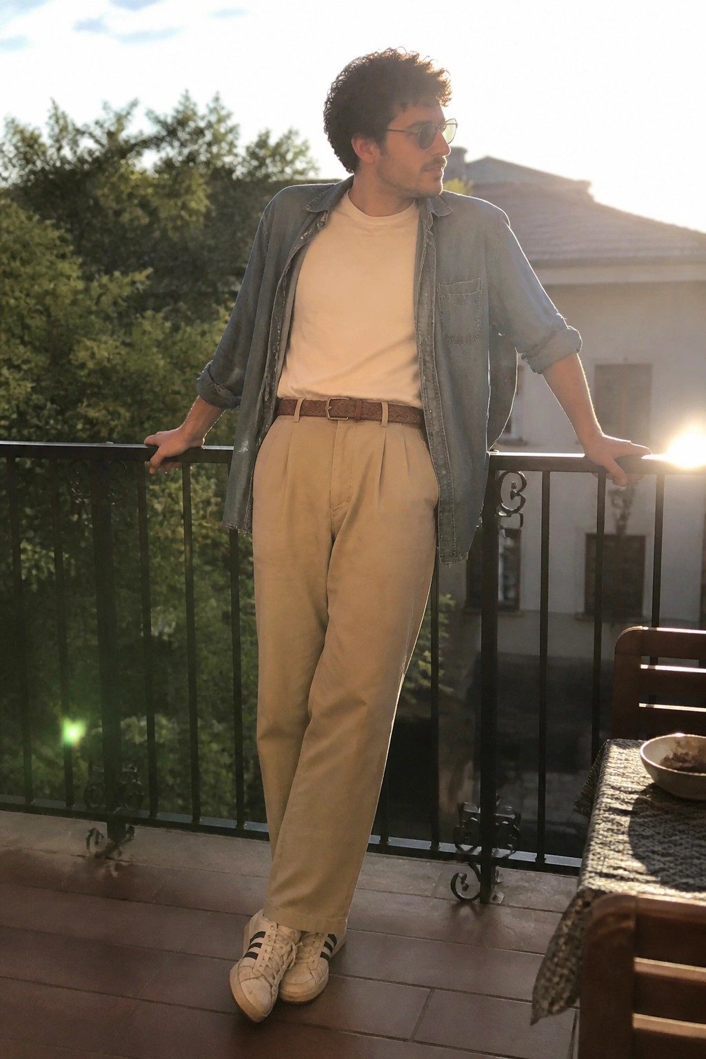 Man in denim shirt, white tee, and pleated khaki pants on sunny balcony railing