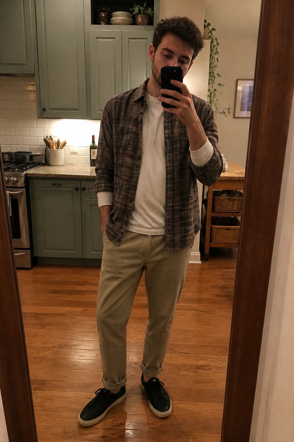 Man in plaid shirt and khakis taking mirror selfie in green kitchen.