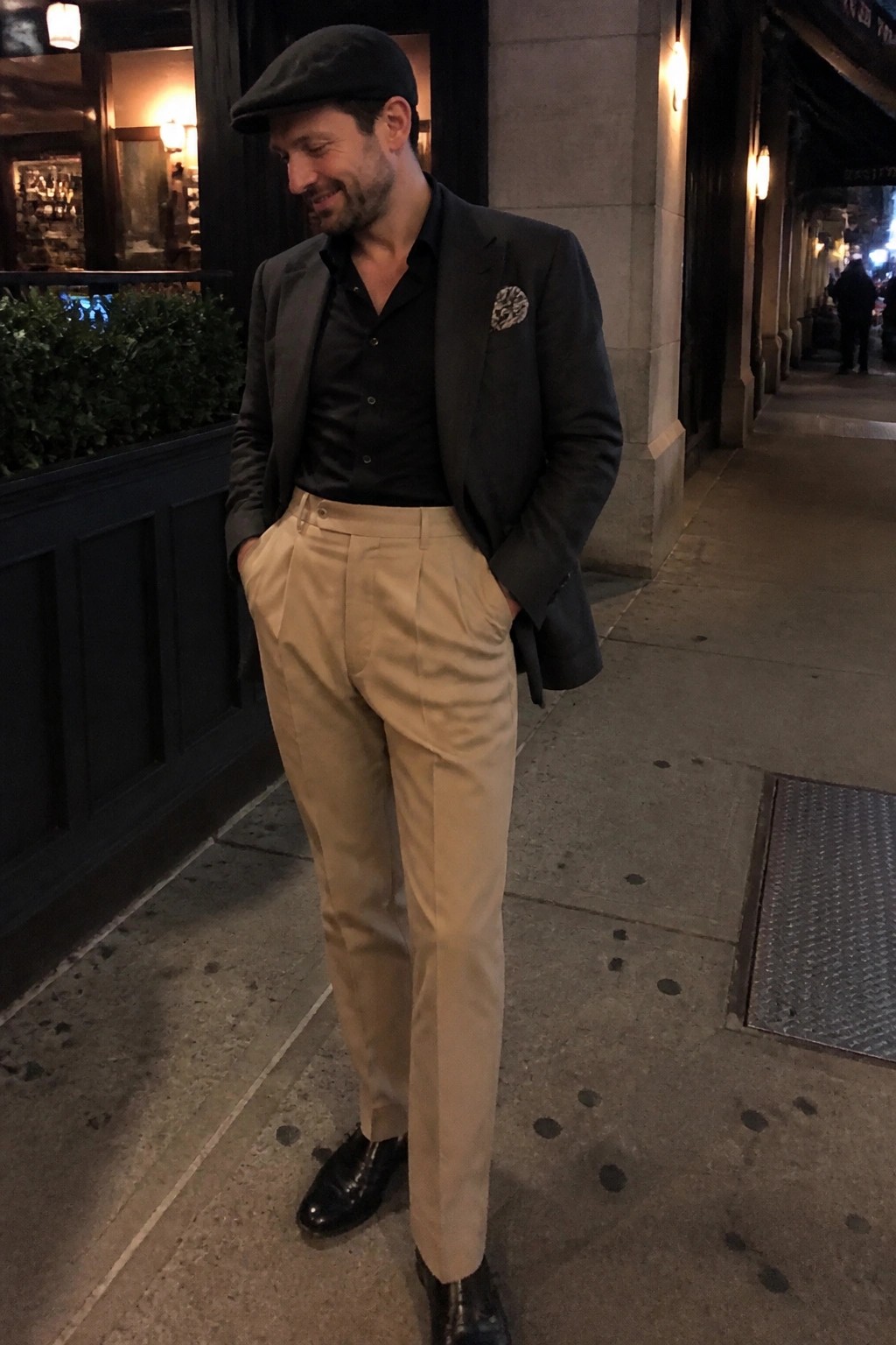 Man in black cap, blazer, and beige trousers on nighttime city sidewalk