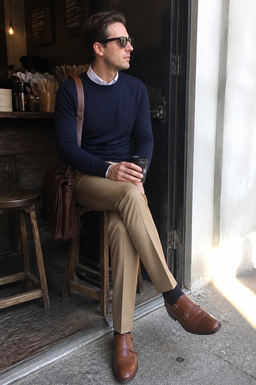 Man in navy sweater and tan pants sits outdoors holding coffee, wearing sunglasses and bag.