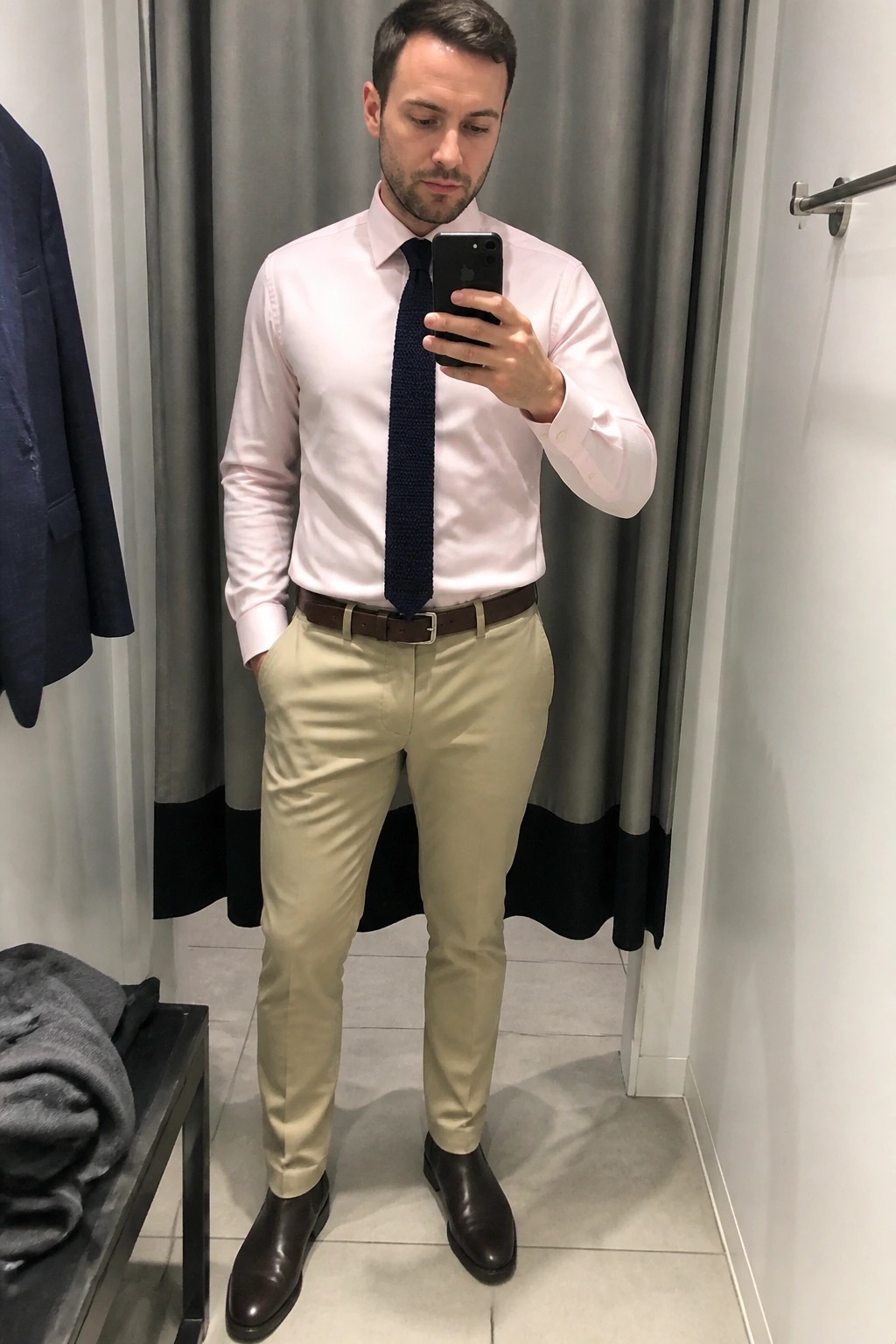 Man in pink shirt with navy tie and khaki pants taking mirror selfie in fitting room