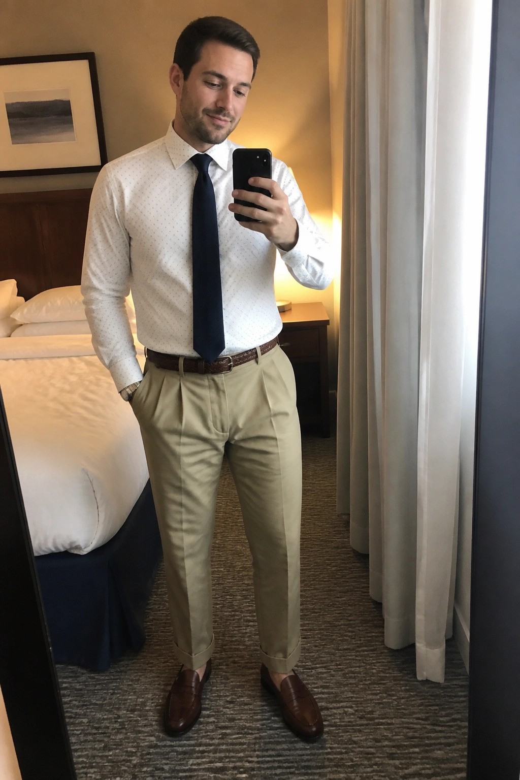 Man in dotted white shirt, navy tie, and khaki pants taking mirror selfie